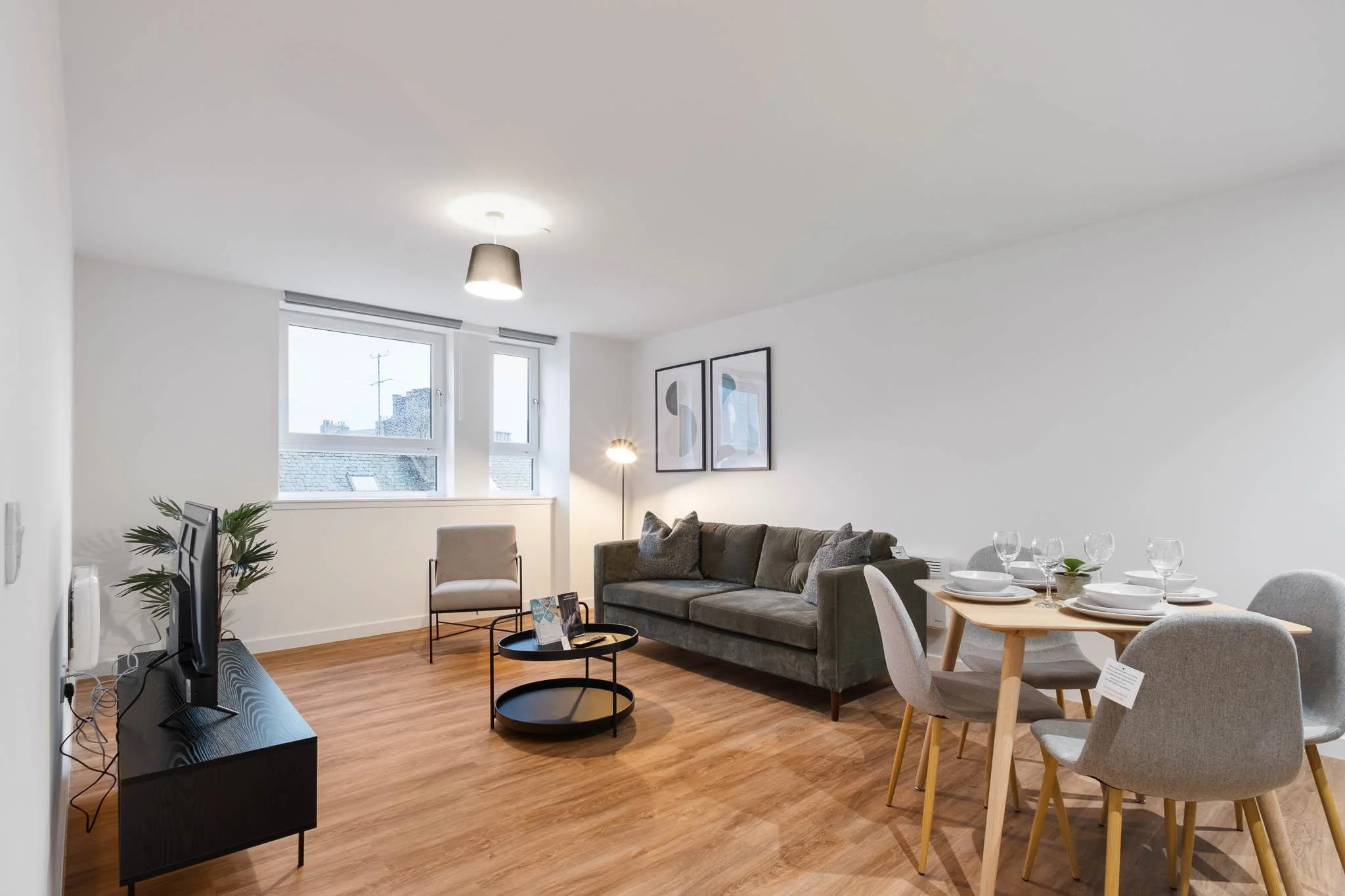 A modern living room with a gray couch, a beige armchair, a large window, and wall art. A dining table with six chairs and tableware is visible on the right. Wooden flooring and a minimalist design.