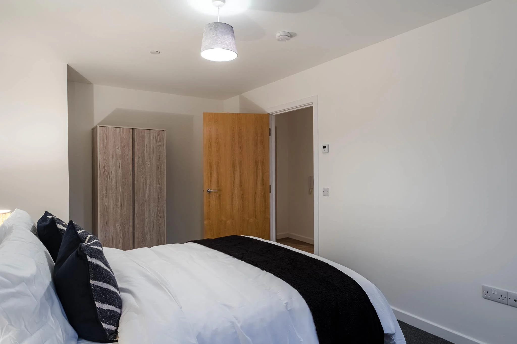 A bedroom with a white bed, black and striped pillows, a wooden wardrobe, an open wooden door, and a plain white wall with a thermostat and power outlet.