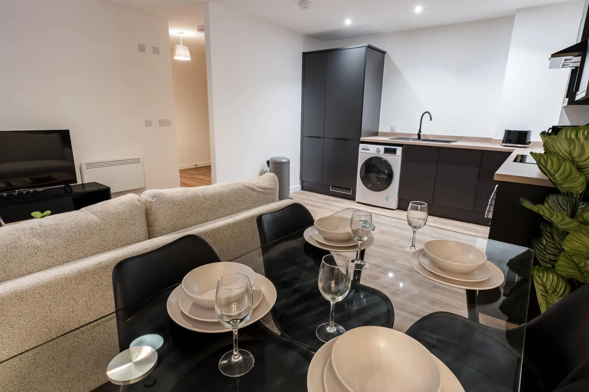 Modern open-plan living space with a kitchen and dining area. The kitchen features dark cabinets, a washing machine, and a black cabinet, with a sink and small appliances on the countertop. The dining area has a black table with black chairs, set wit