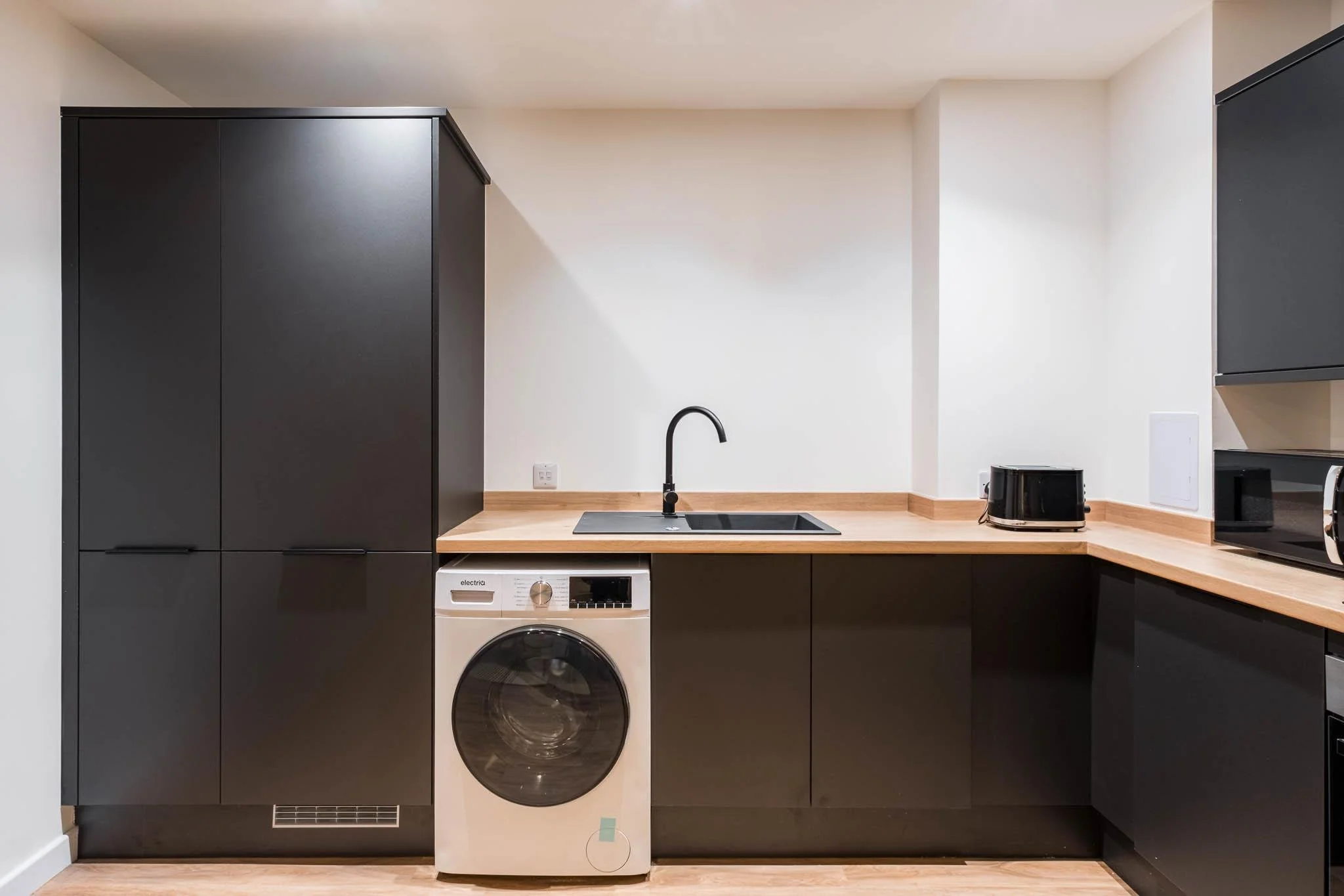 Modern kitchen with black cabinets, a wooden countertop, a black faucet, and appliances including a washing machine, toaster, and microwave.