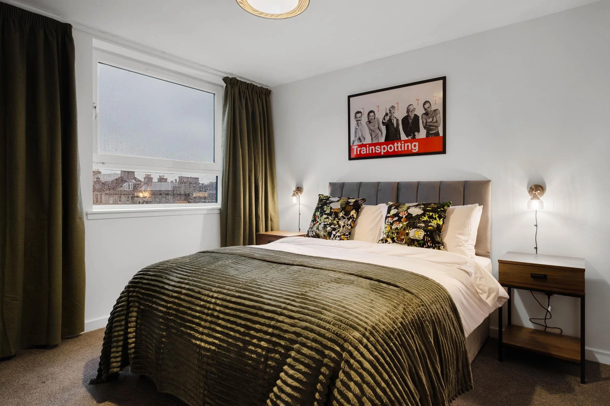 A cozy bedroom with a large window covered by dark green curtains, a bed with a textured green bedspread, patterned pillows, and a picture on the wall with the word 'Trainspotting' and images of five people.