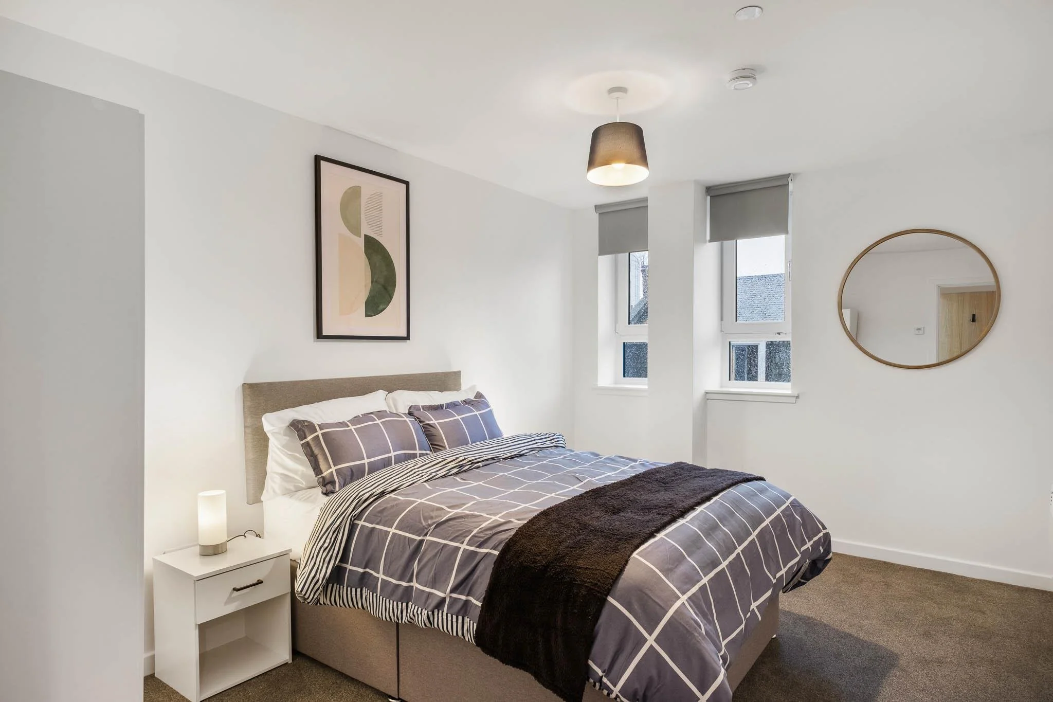 A modern bedroom with a bed that has a beige headboard, checkered bedding, and a dark brown throw blanket. There is a white nightstand with a white lamp beside the bed. The walls are white, decorated with minimalist artwork and a round mirror. Natura
