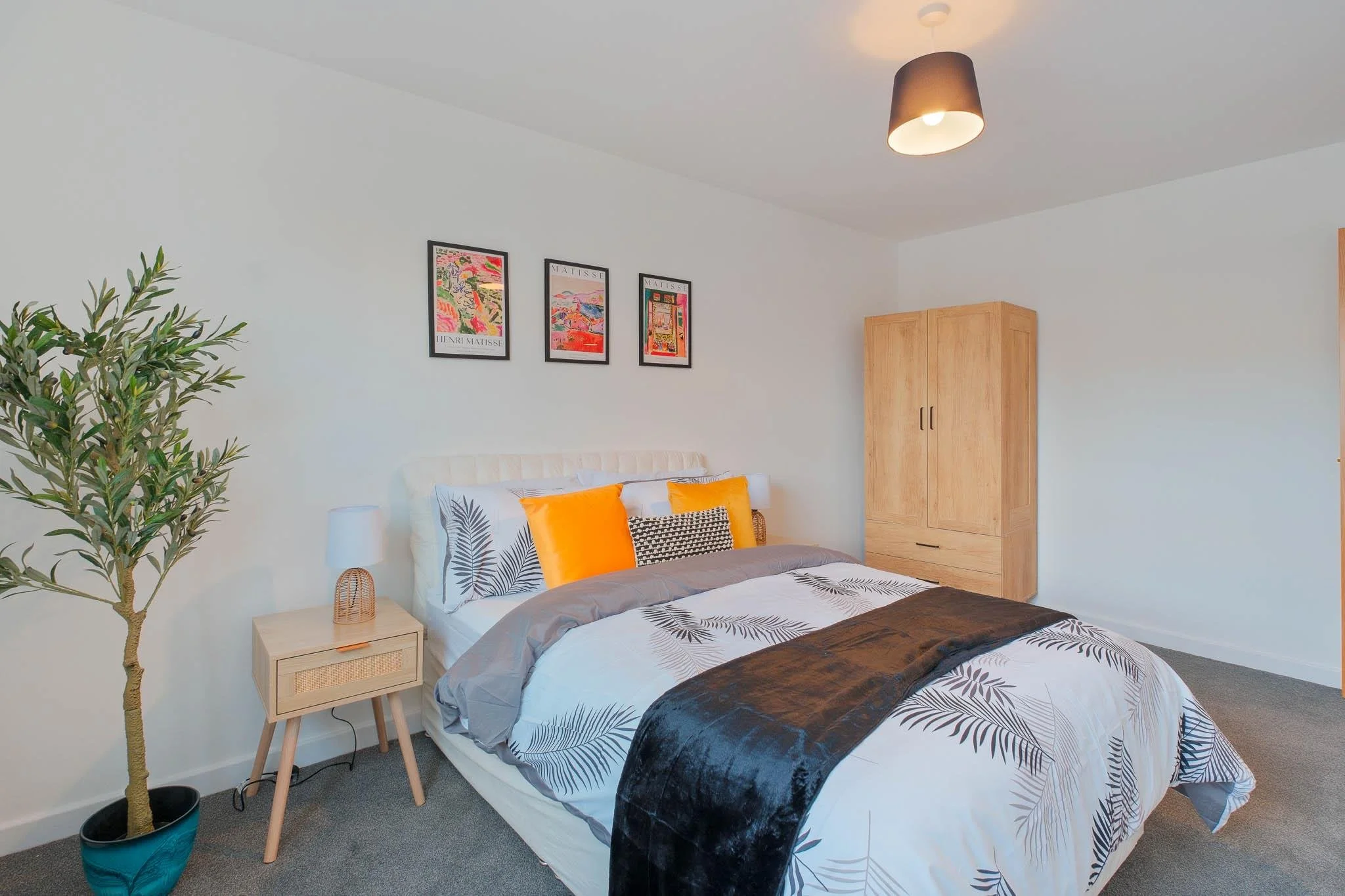 A bedroom with white walls and a bed with black, white, and orange bedding. There is a small wooden nightstand with a lamp, a tall wooden wardrobe, framed colorful artwork on the wall, a potted plant in the corner, and a ceiling light fixture.