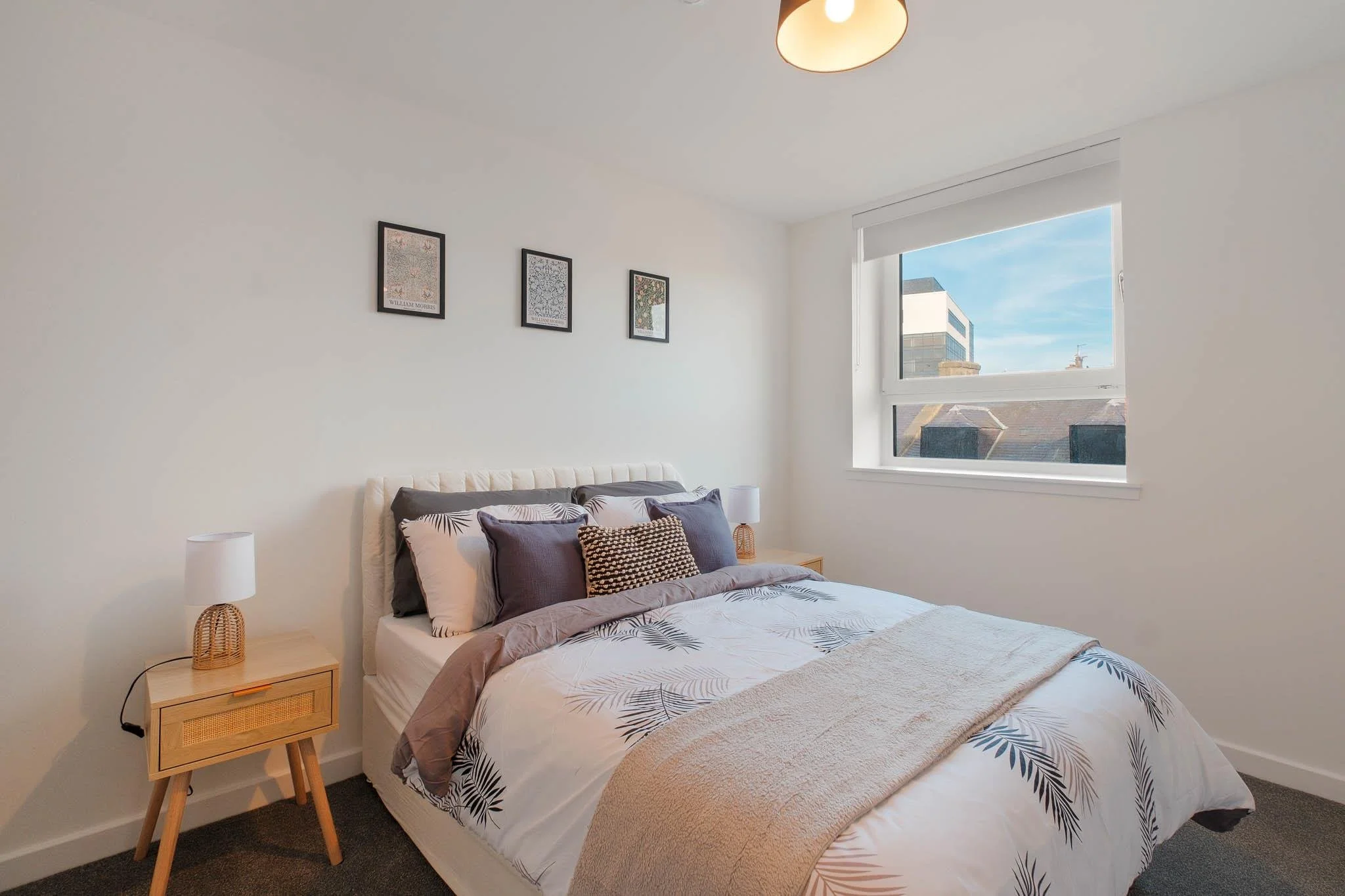 A bedroom with a bed that has patterned pillows and bedding, a wooden bedside table with a white lamp, a window with a view of neighboring rooftops and a clear sky, and three framed art prints on the white wall.