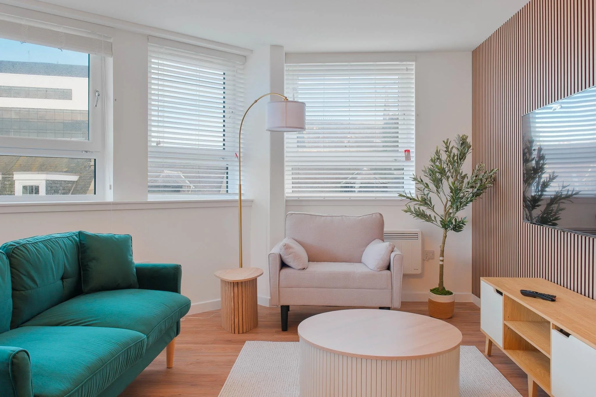 Living room with large windows with white blinds, a green sofa, a beige armchair with small pillows, a wooden side table, a tall floor lamp, a potted plant, a white round coffee table, and a wooden TV stand. There is a wood-paneled accent wall and a 