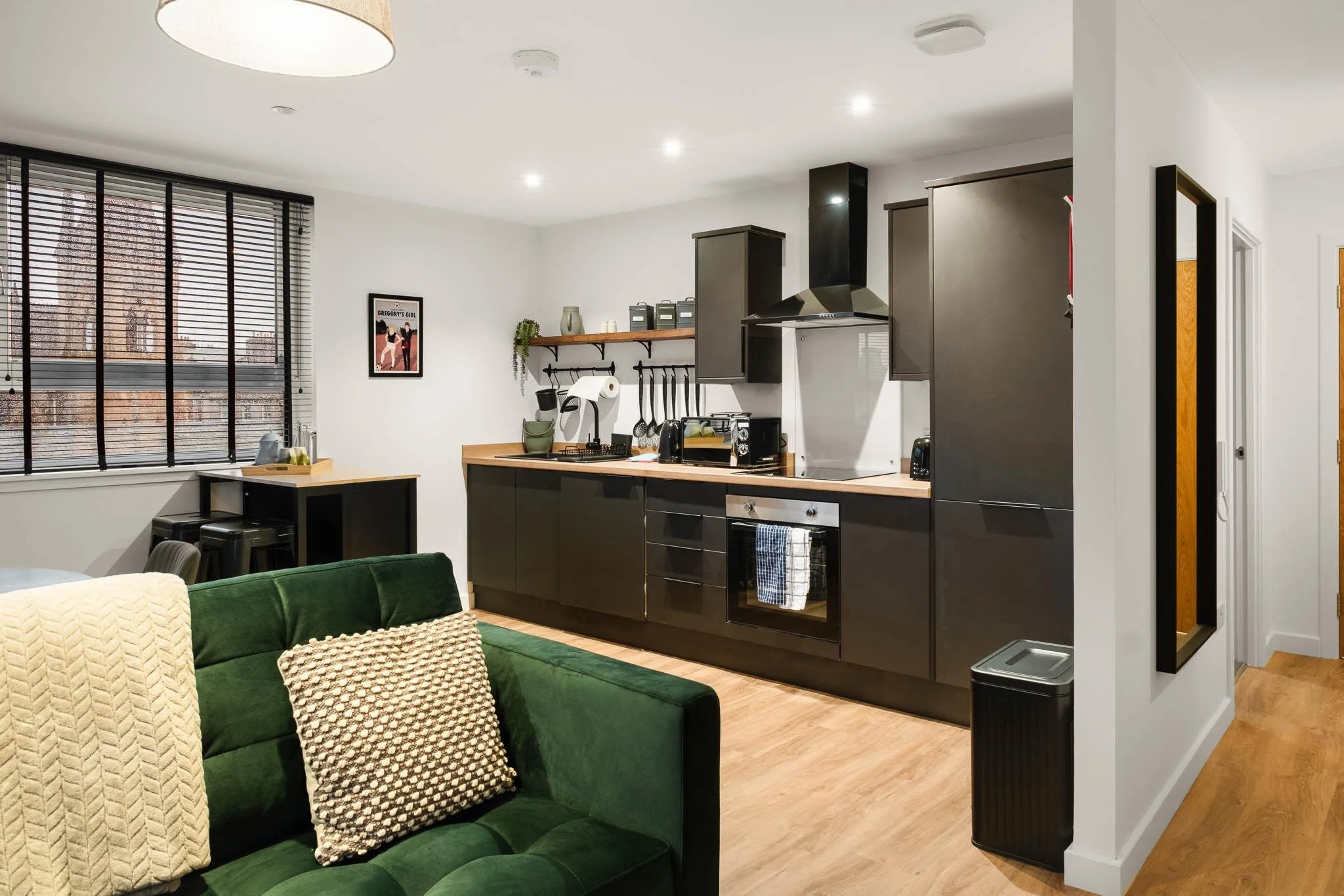 Modern kitchen with black cabinets, wooden countertops, and open shelving. A window with black blinds lets in natural light. In the foreground, a green sofa with beige and patterned pillows is visible.