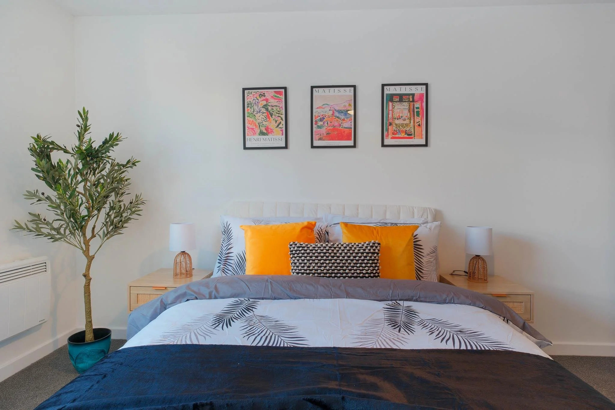 A bedroom with a bed that has black and white patterned pillows and orange pillows, a gray blanket, and a leaf-printed bedsheet. There is a picture of three framed colorful abstract artworks labeled 'Matisse' and 'Henri Matisse' hanging on the white 