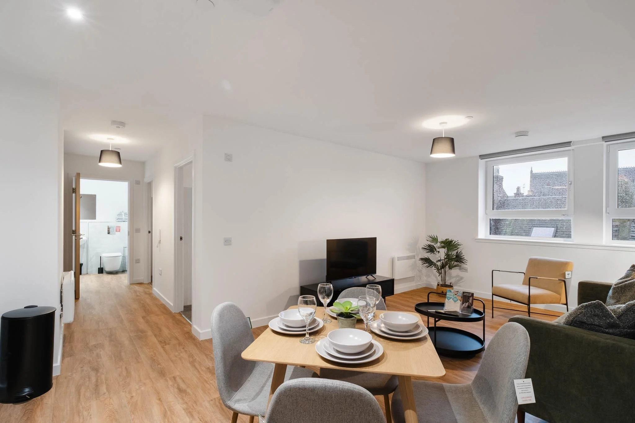 Interior of a modern apartment with a dining table set with white dishes and glasses, an arrangement of gray chairs, a living area with a TV, sofa, armchair, and a window showing rooftops outside.