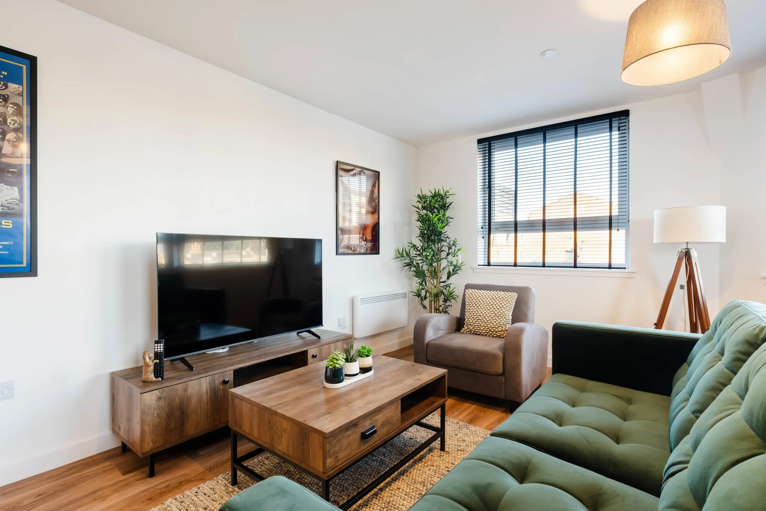 Living room with white walls, large window with blinds, a green sofa, beige armchair with a patterned pillow, TV on a wooden stand, coffee table with decor, a tall floor lamp, and houseplants.
