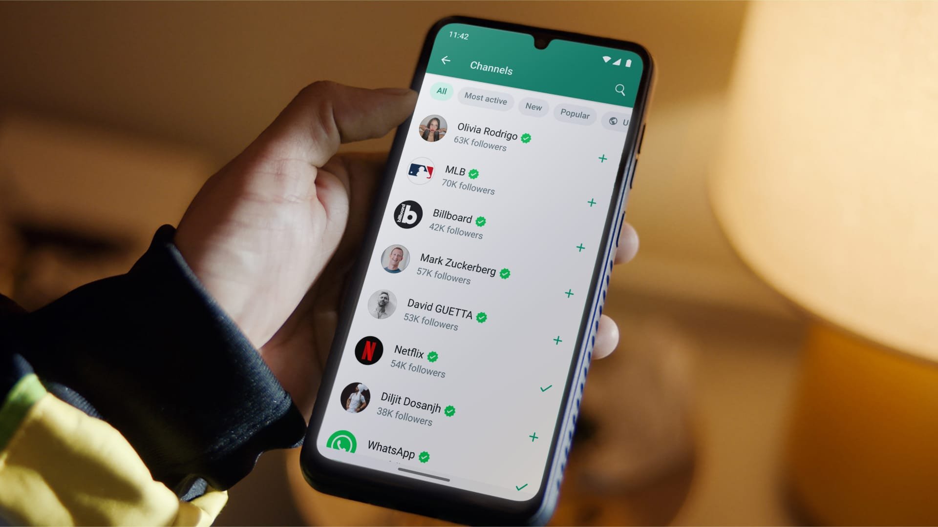A person holding a smartphone displaying a WhatsApp Channels with a list of channels and verified checkmarks.