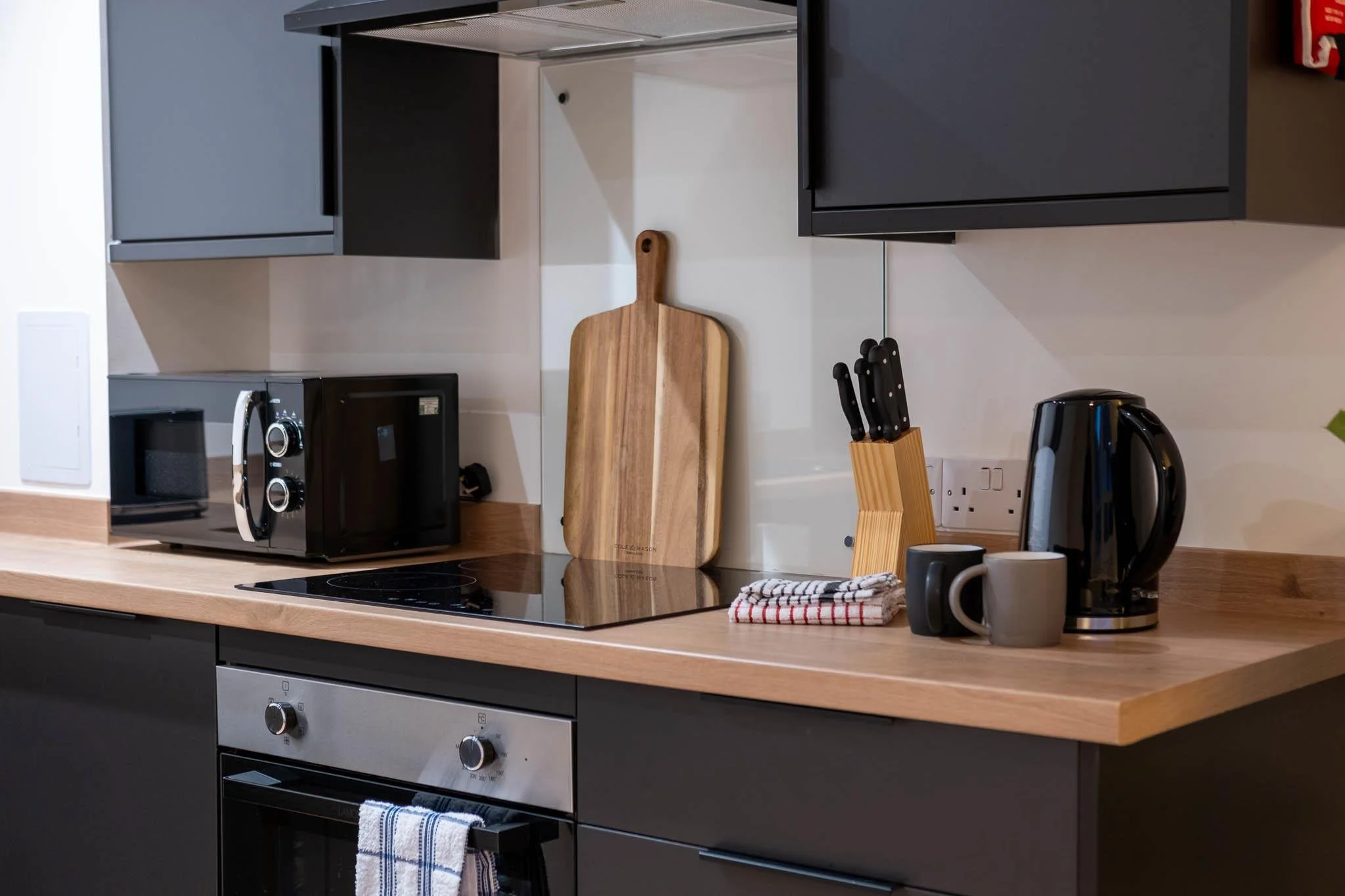 Kitchen countertop with a microwave, a wooden cutting board, a knife block with knives, an electric kettle, mugs, a dish towel, and black cabinets with upper cabinets and a stove below.