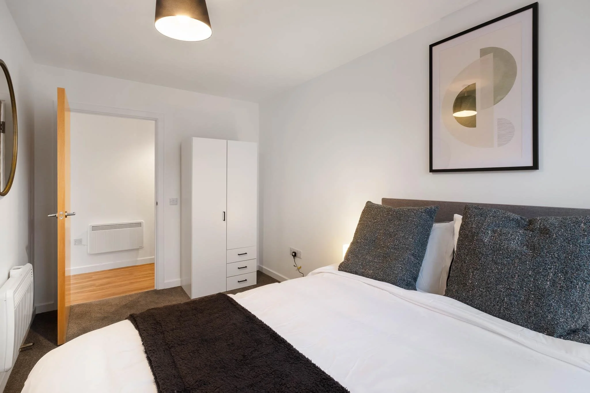 A modern bedroom with a white wall, framed abstract art, a bed with white sheets and dark gray pillows, a dark brown rug, a white wardrobe, and a partly open wooden door revealing a room with wooden flooring.