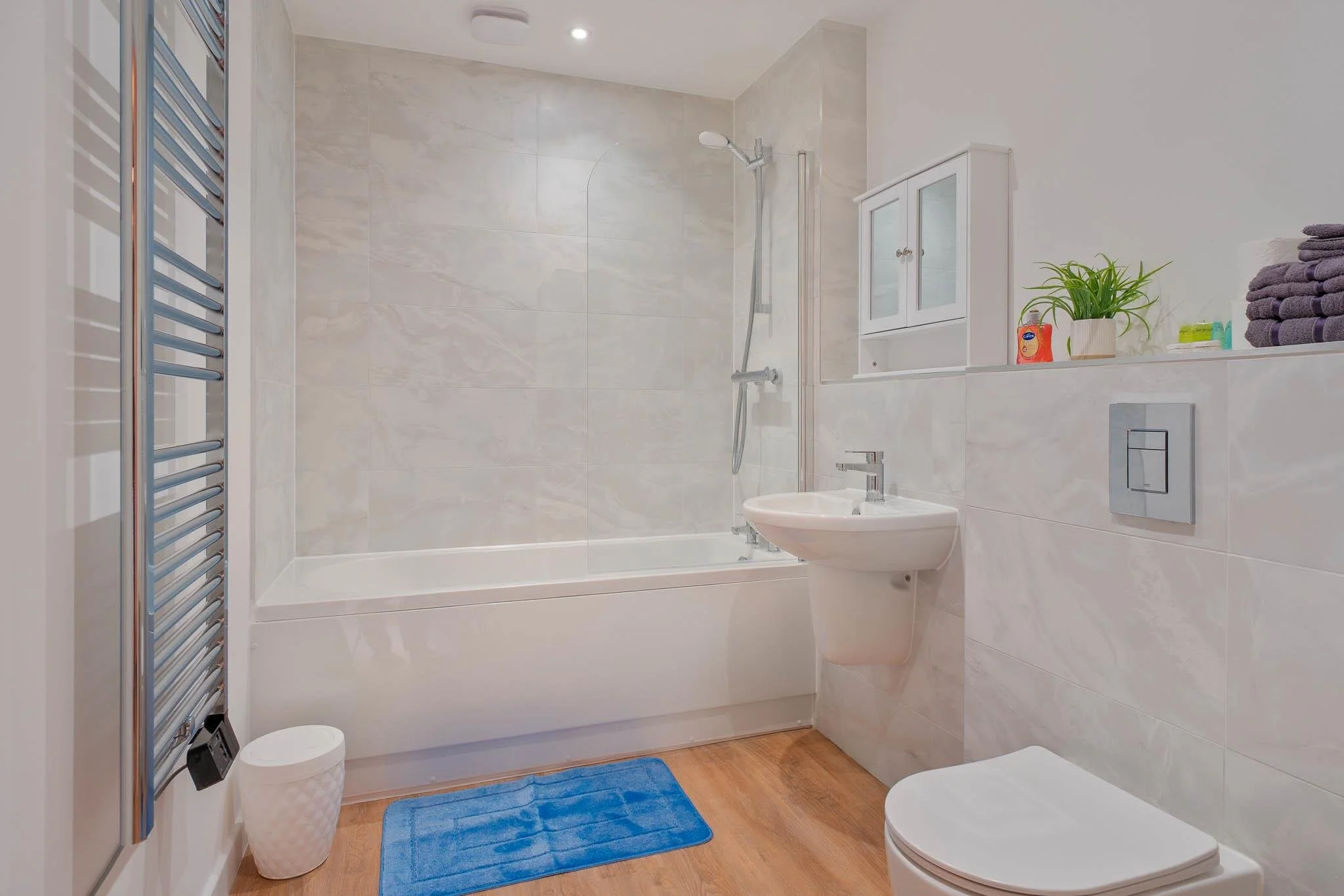 A modern bathroom with a bathtub and shower, a bidet, a wall-mounted toilet, a cabinet with glass doors, towels, a potted plant, and a towel warmer on the left wall. There is a blue bath mat on the wooden floor.