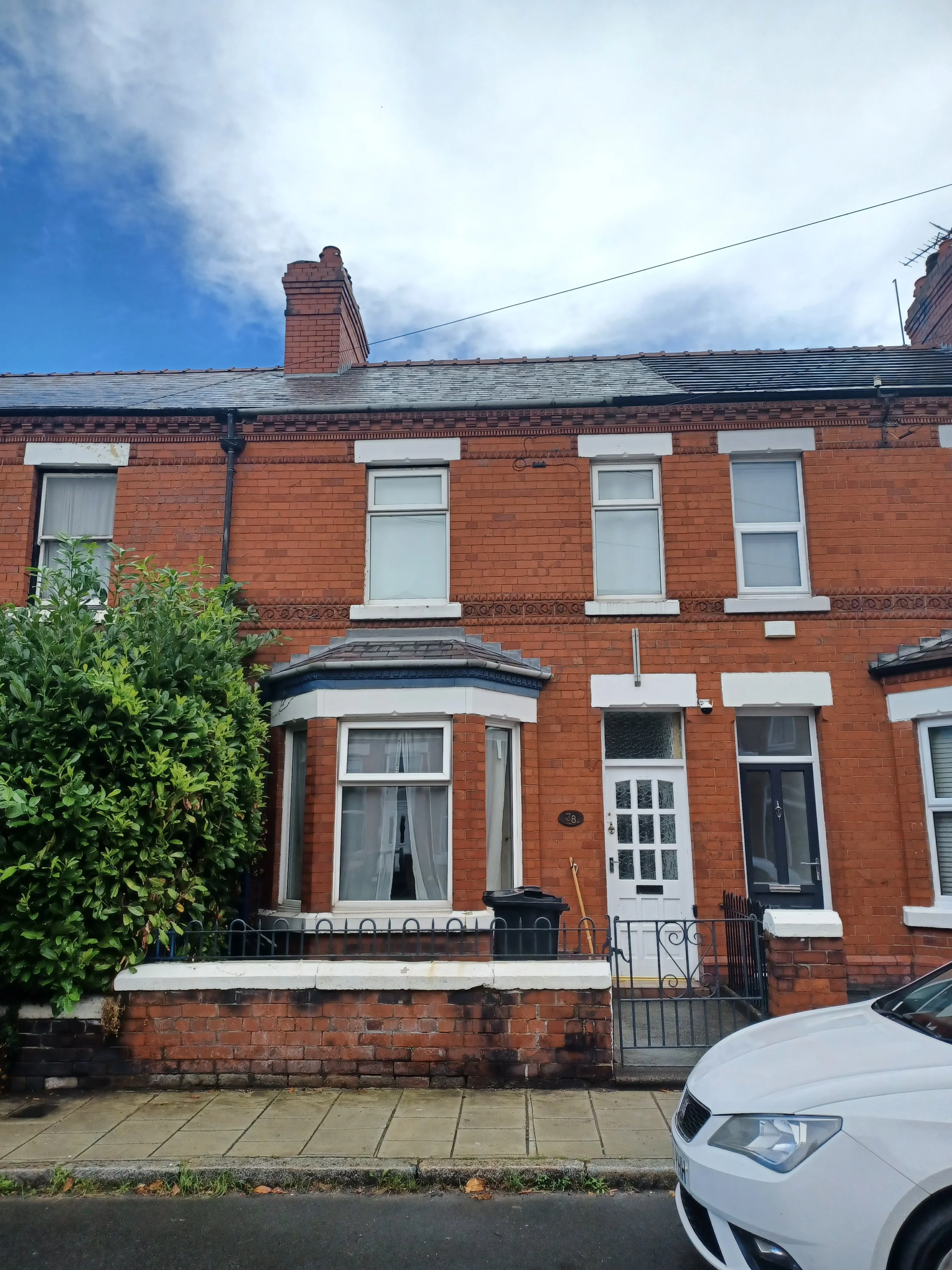 Red brick Victorian terrace house with white window frames and a small front yard. There are three large windows on the upper floor and a bay window on the ground floor. A white security door and a black gate enclose the front yard. A green shrub is on the left side of the house, and a white car is parked in front on the street.