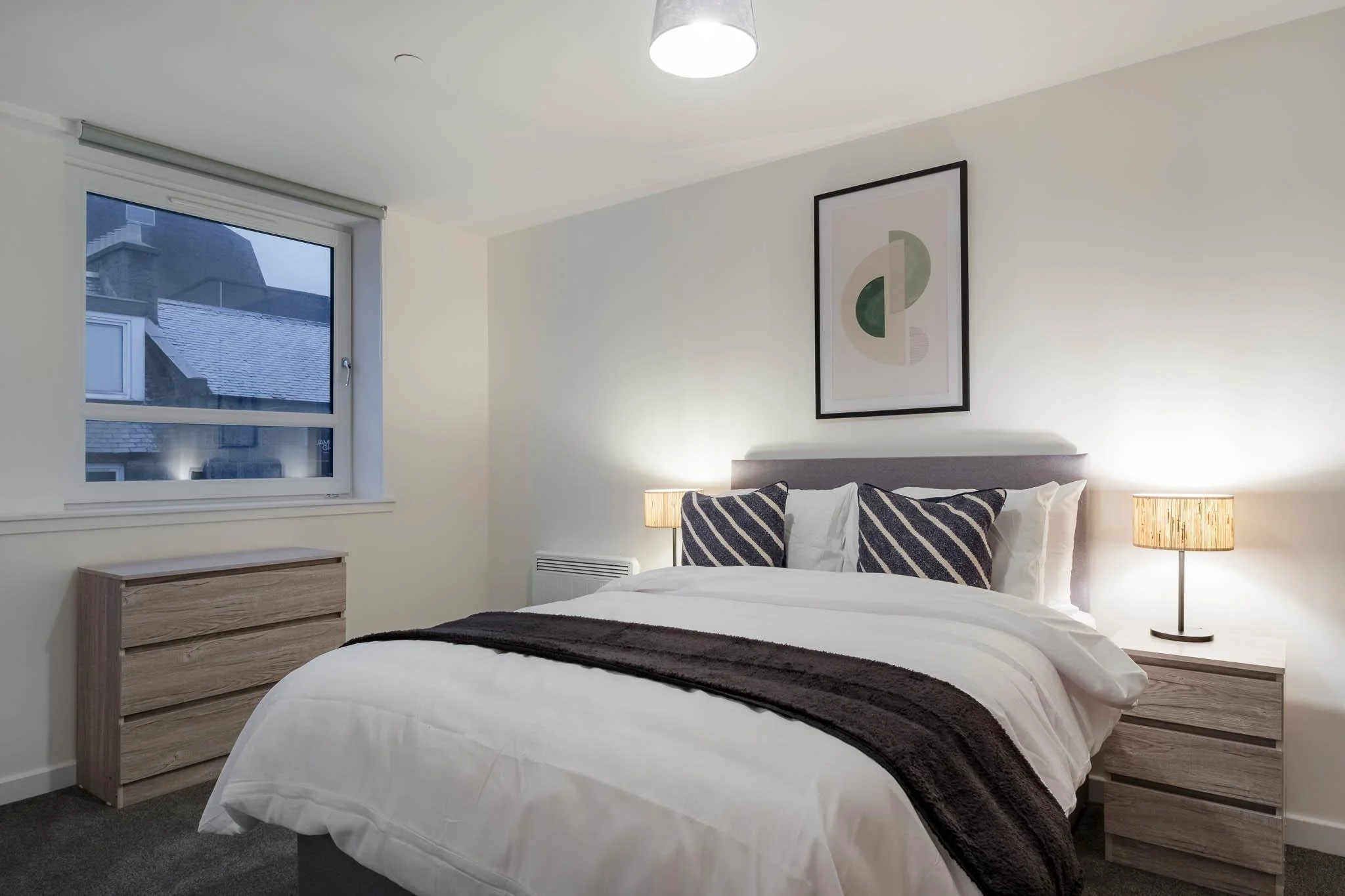 A modern bedroom with a large bed, two matching wooden bedside tables with lamps, a window looking out on rooftops, and minimalist wall art. The room has a ceiling light, white walls, and a cozy atmosphere.