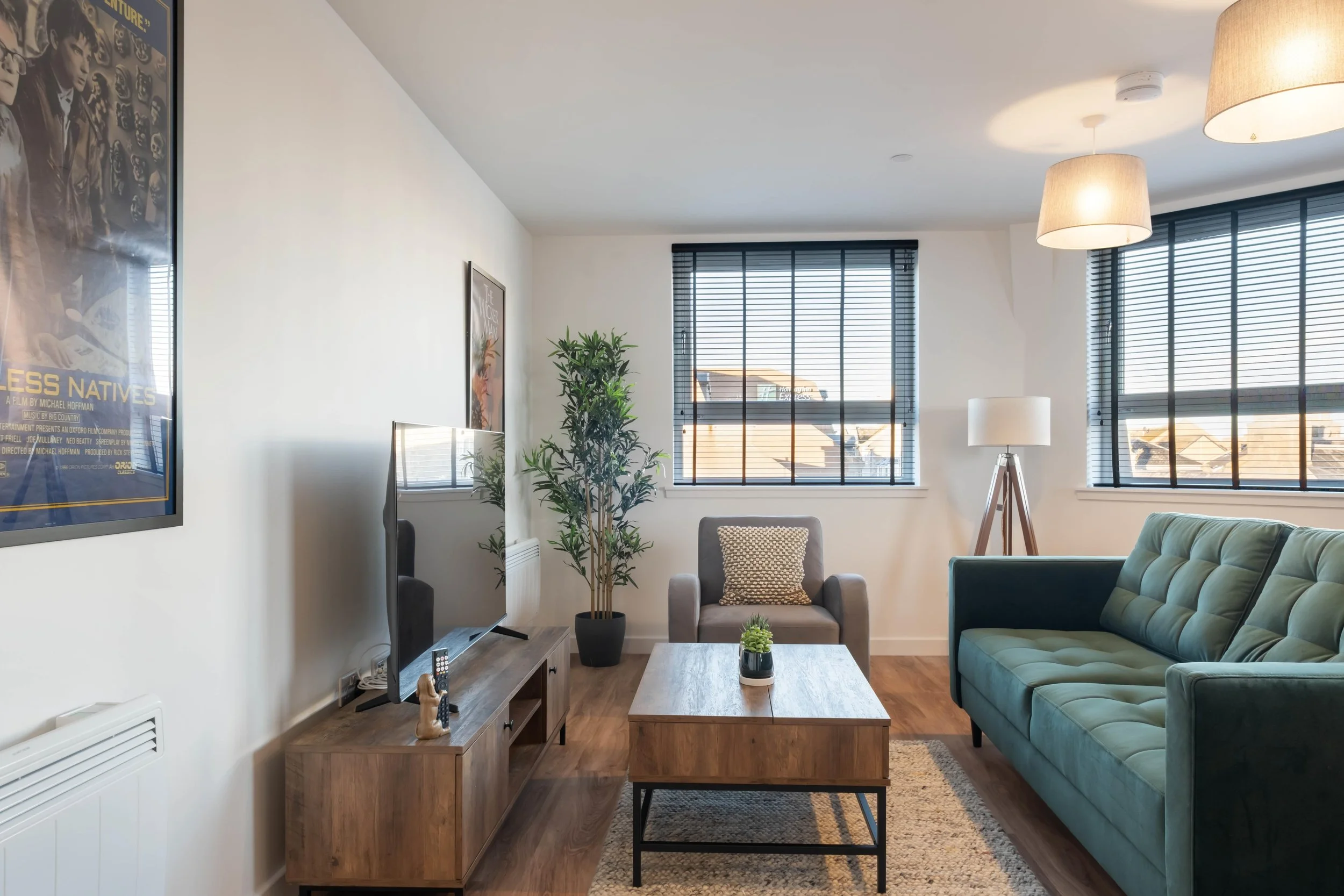 A modern living room with large windows, black blinds, a teal sofa, a single armchair with a geometric pillow, a wooden coffee table, a tall potted plant, a TV on a wooden stand, a floor lamp, two ceiling pendant lights, and framed artwork on the walls.