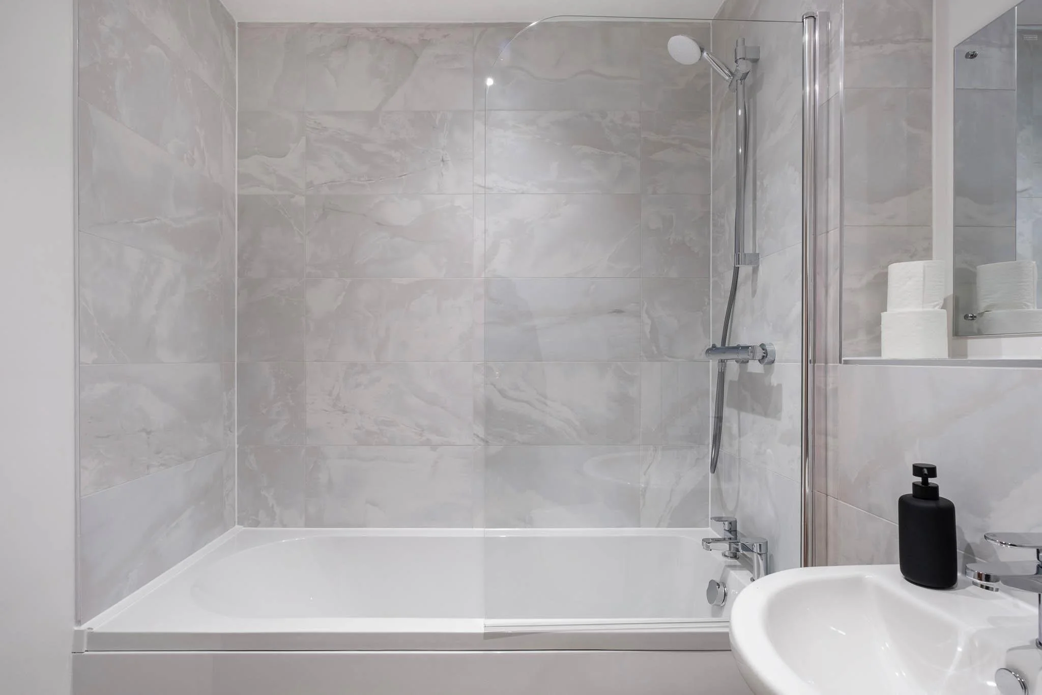 A modern bathroom shower with large gray marble tiles, a clear glass shower door, a handheld shower head with a chrome finish, a small sink with a black soap dispenser, and a mirror with toilet paper rolls reflection.