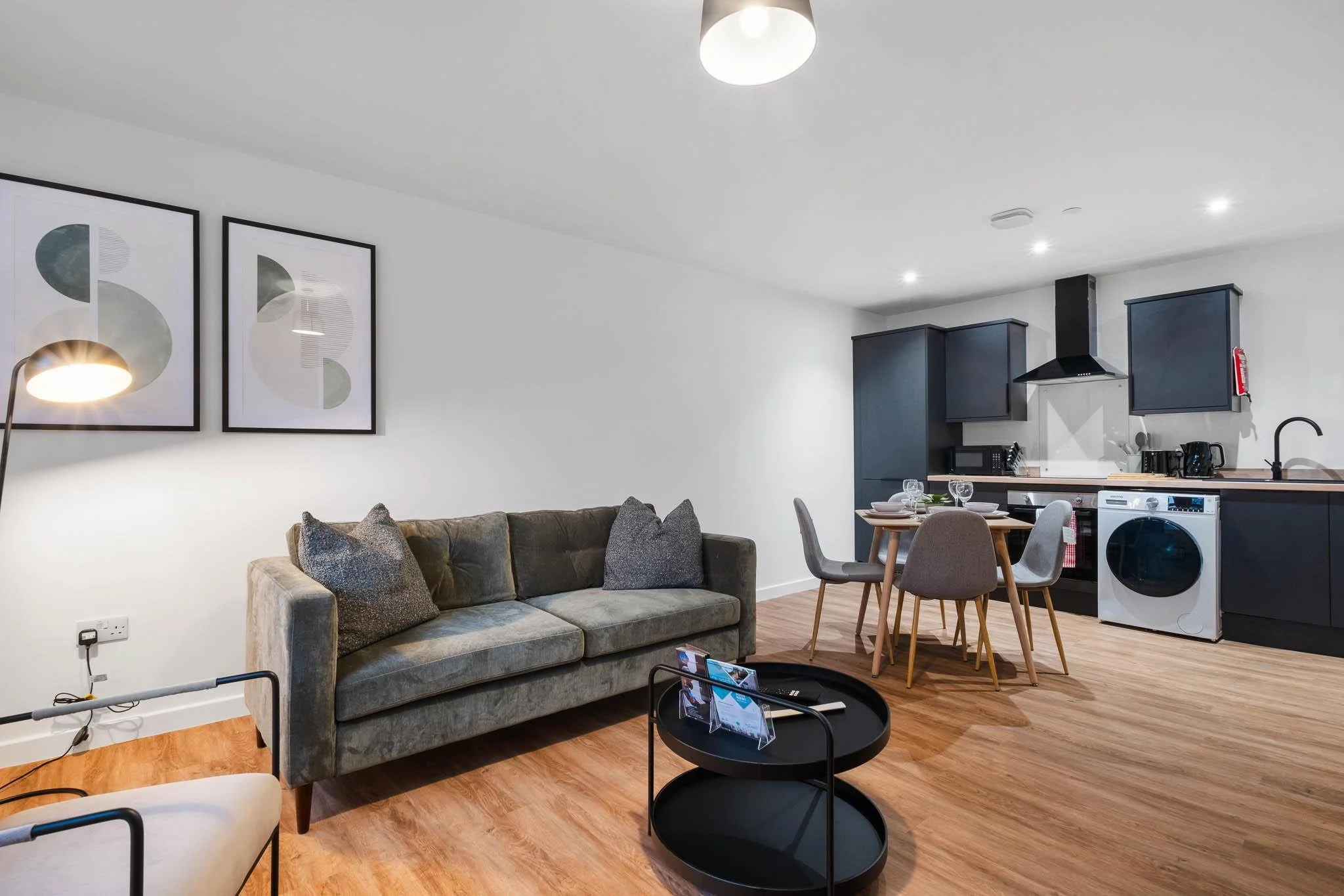 A modern, open-plan living room and kitchen with minimalist decor. The living room has a gray sofa with textured pillows, abstract framed art on the wall, and a floor lamp. The kitchen features dark cabinets, white walls, a stainless steel range hood