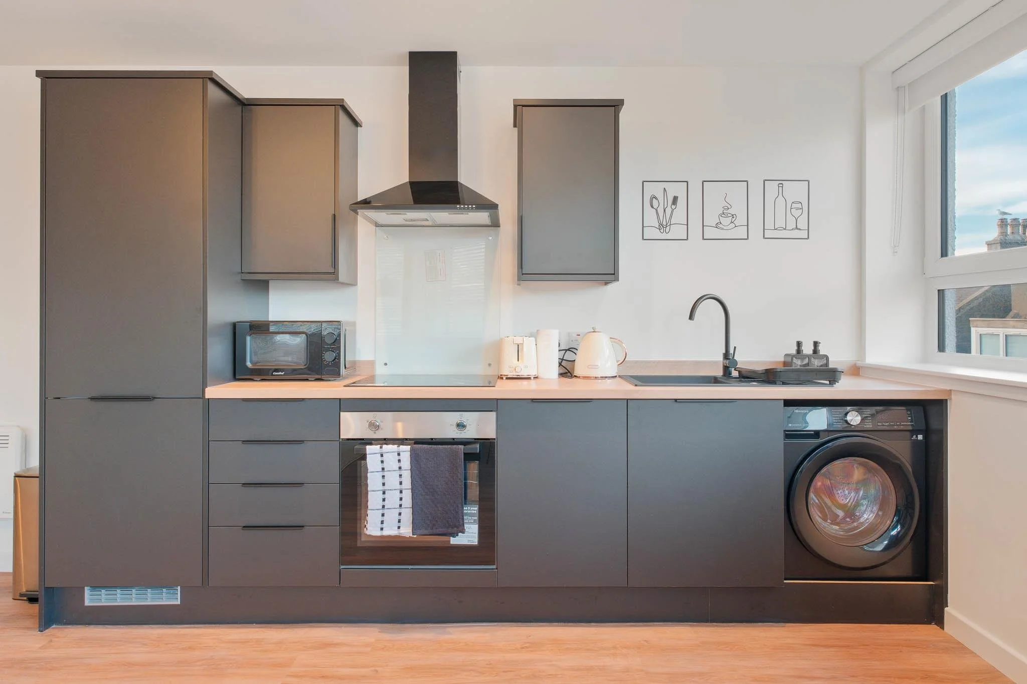 Modern kitchen with dark gray cabinets, light-colored countertop, appliances including oven, microwave, kettle, and washing machine, and wall art of line drawings of cutlery, coffee, and wine glass near a large window.