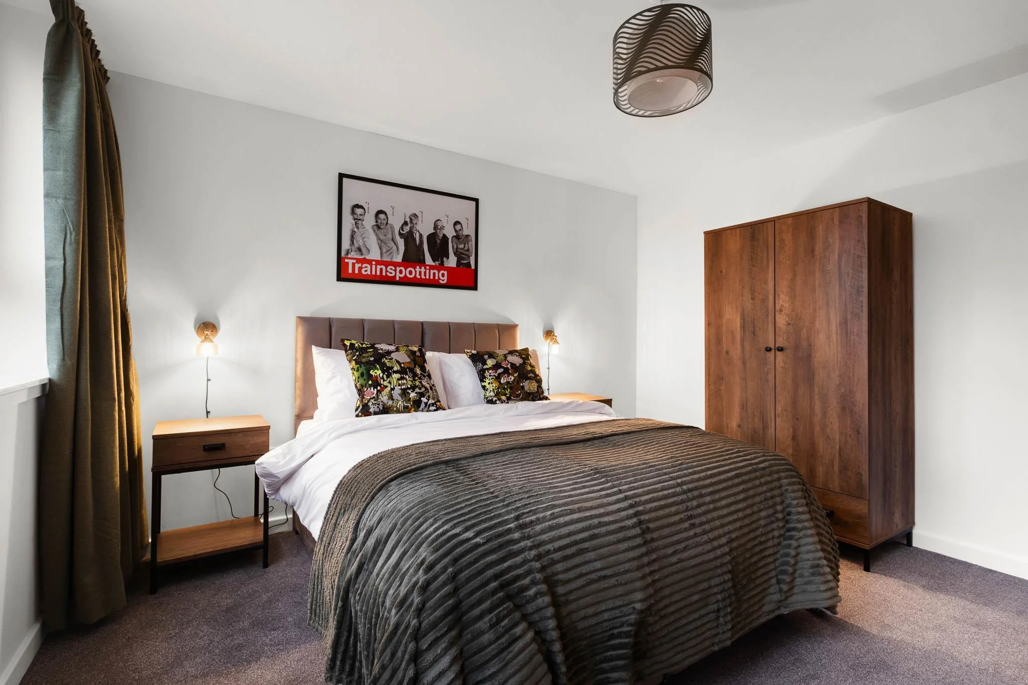 A bedroom with a bed that has a beige headboard, striped bedding, and decorative pillows. There is a wooden wardrobe on the right, bedside tables with lamps, a window with green curtains, a modern ceiling light, and a framed black-and-white photo of 