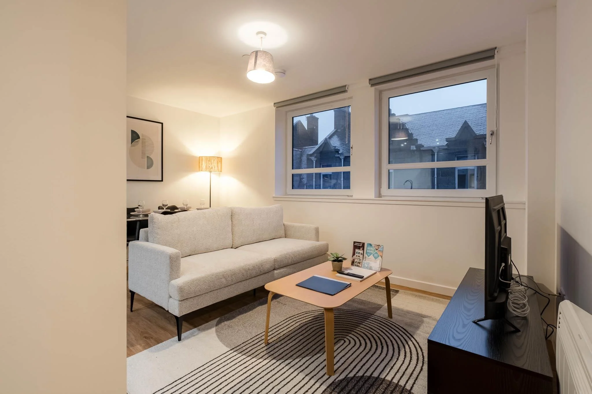 Living room with a white sofa, wooden coffee table, black entertainment center with a television, striped rug, large windows, artwork on the wall, and a floor lamp, during dusk.