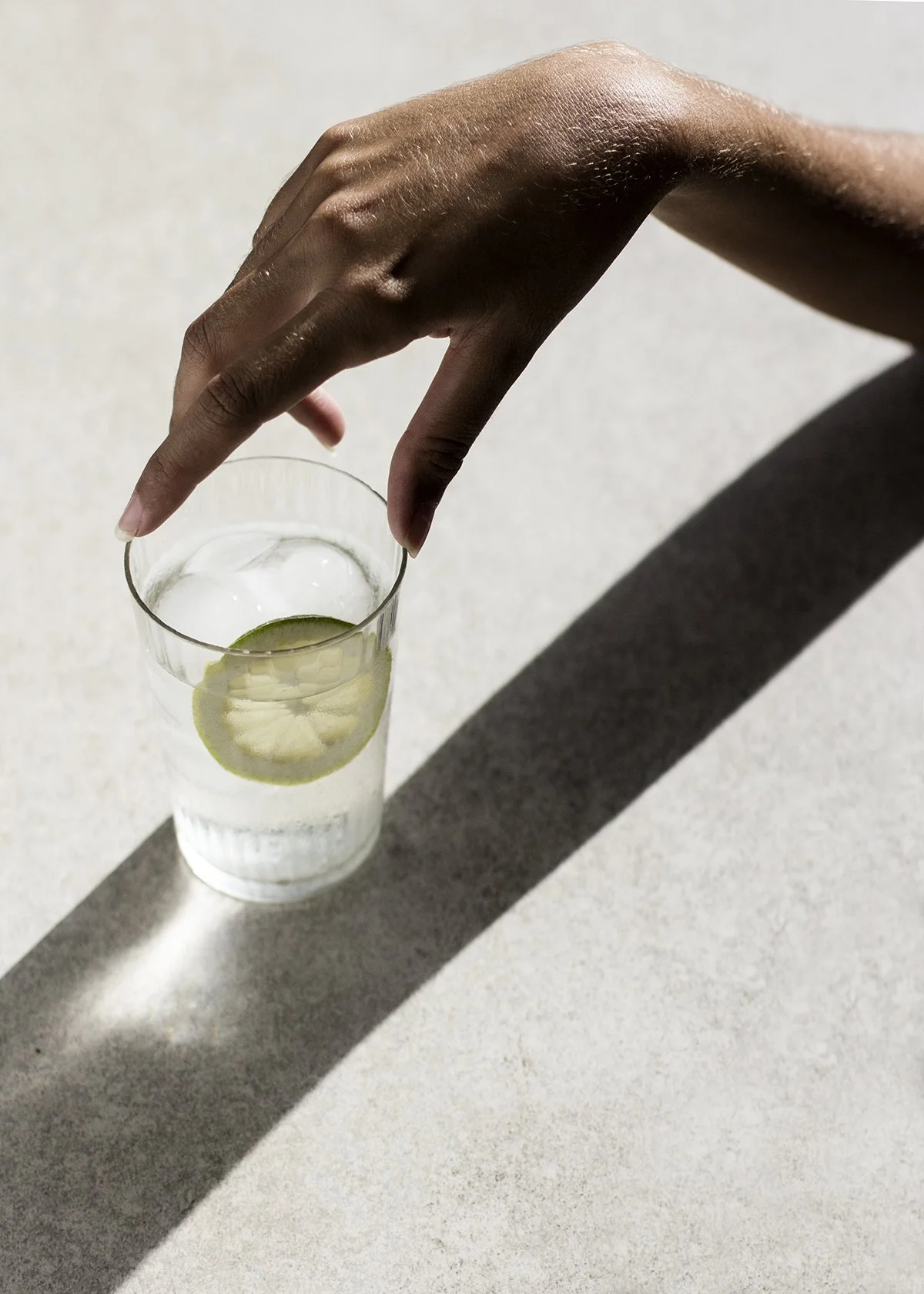 Una mano sostiene un vaso con agua y una rodaja de limón, en un fondo claro con sombra.