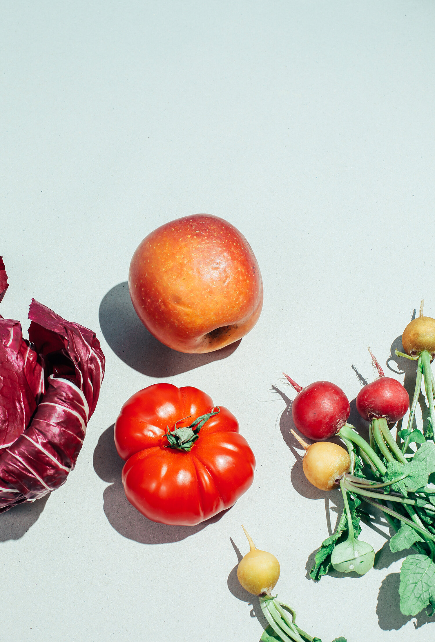 Diversos vegetales y frutas en un fondo claro, incluyendo una pequeña cabeza de lechuga, una manzana, un tomate, rábanos y nabos.