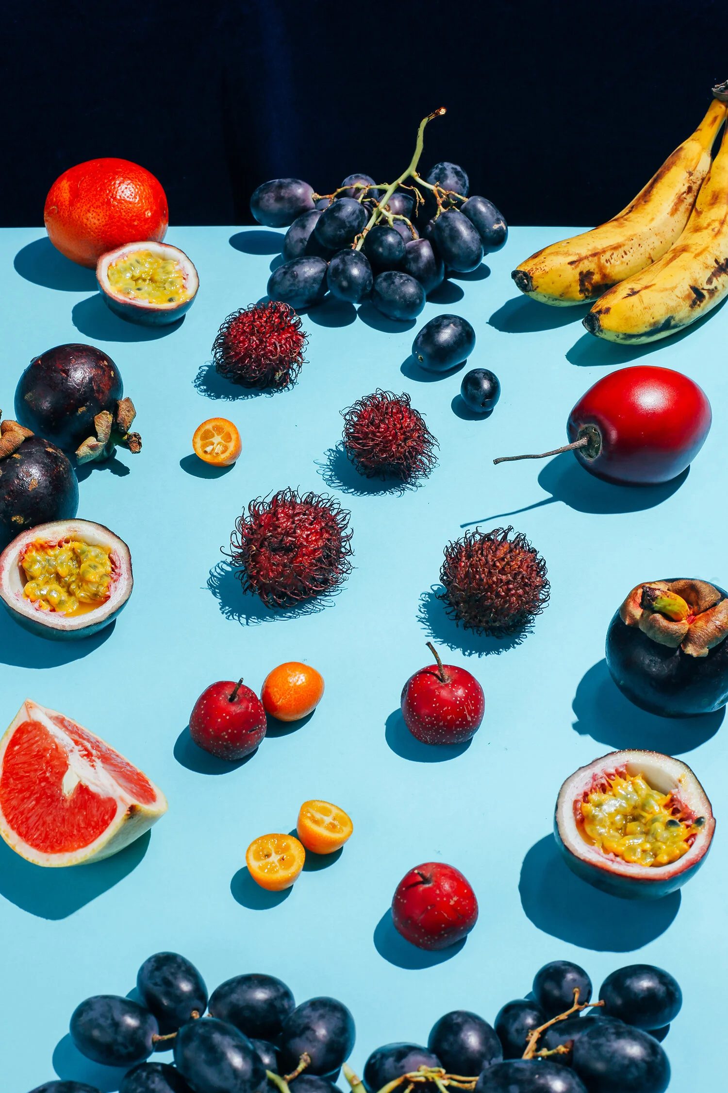 Variedad de frutas tropicales sobre una mesa azul, incluyendo uvas, plátanos, passion fruit, toronjas, manzanas, tomate kumquat, y un aguacate.