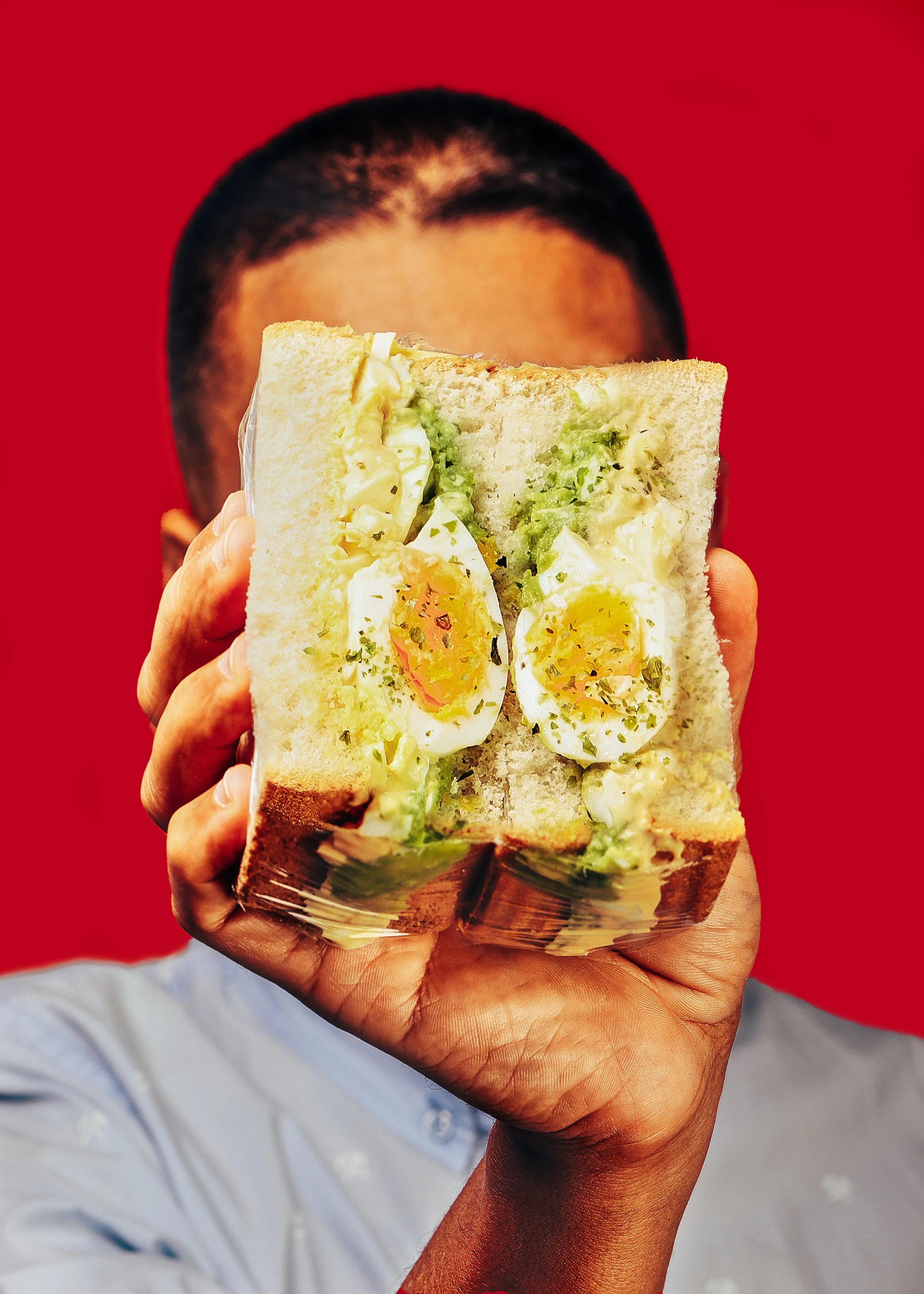 Persona sosteniendo un sándwich japones con huevo cocido, lechuga y mayonesa en un fondo rojo.