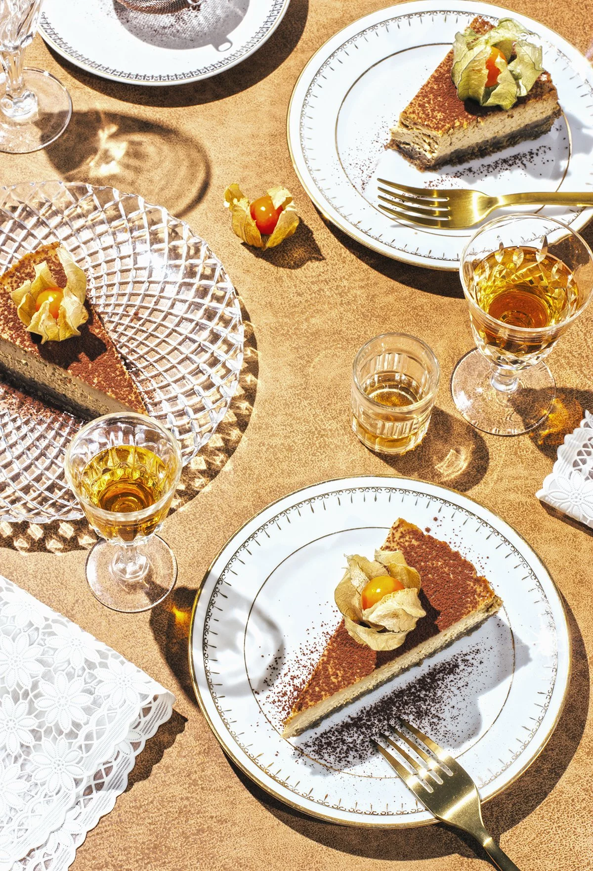 Platos de tiramisú decorados con physalis, copas de vino y vasos de licor en una mesa con manteles de color piel.