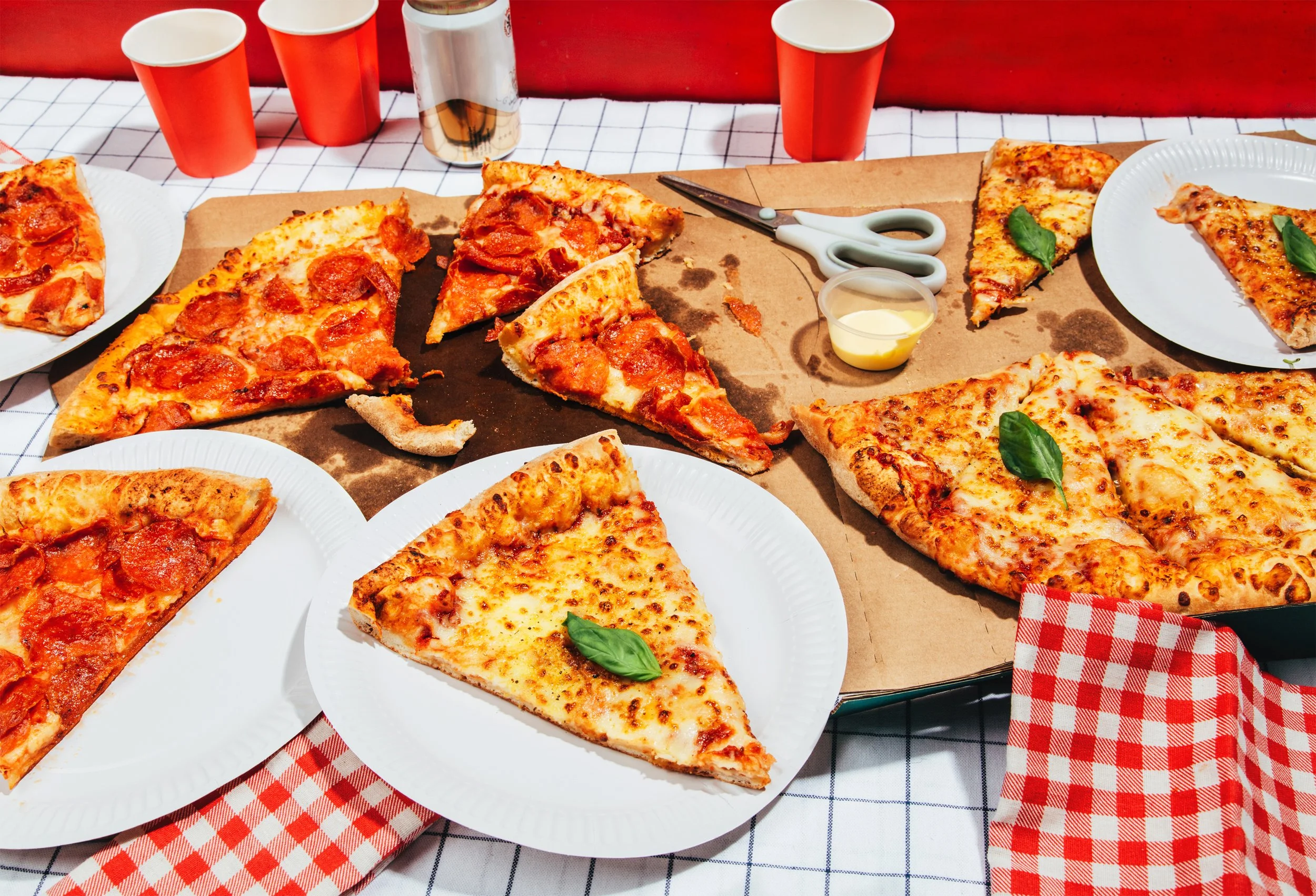 Variedad de porciones de pizza con diferentes ingredientes, algunas decoradas con hojas de albahaca fresca, en una mesa con mantel de cuadros y vasos de plástico rojos.