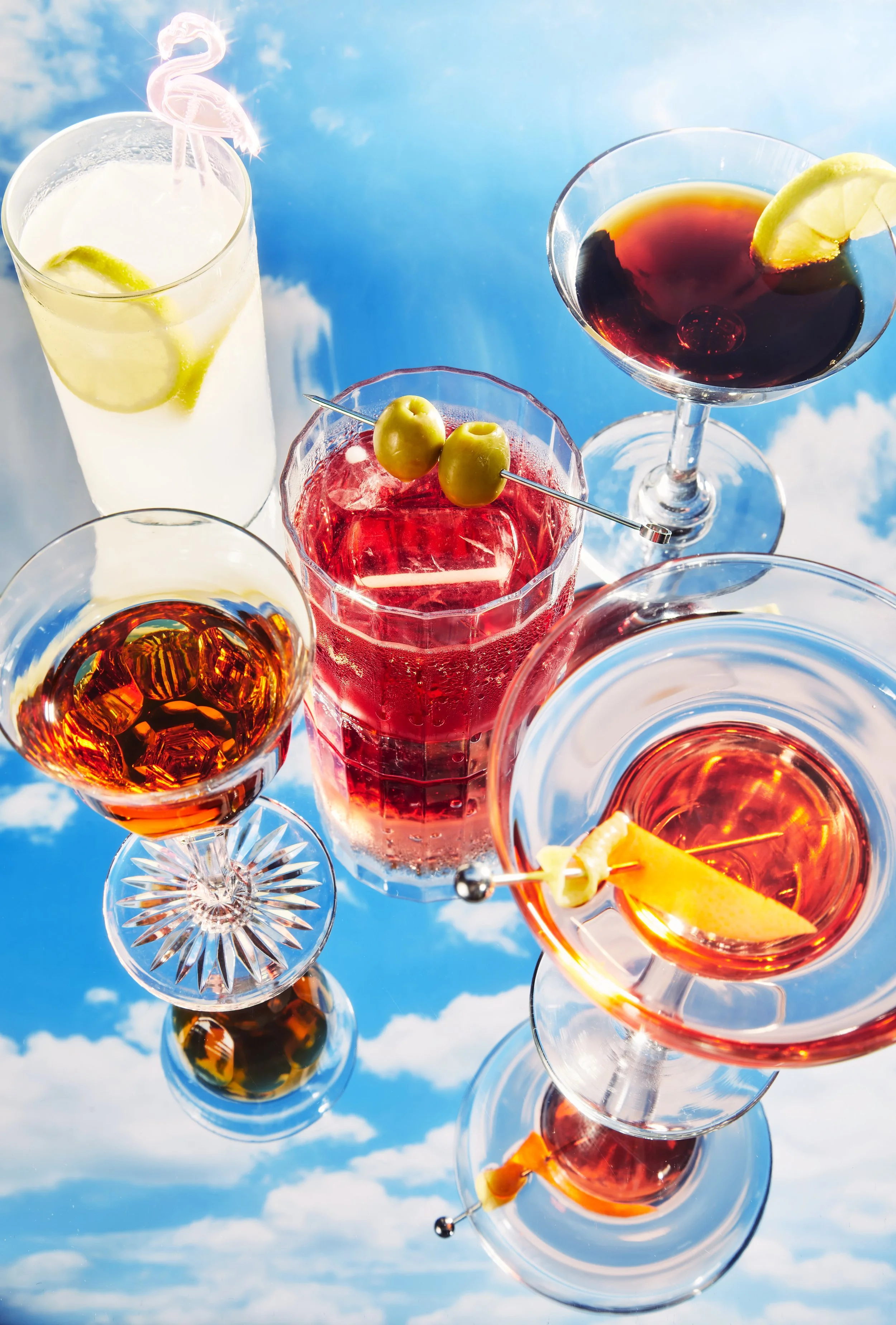 Cinco cócteles con frutas y decoraciones, sobre un fondo de cielo con nubes.
