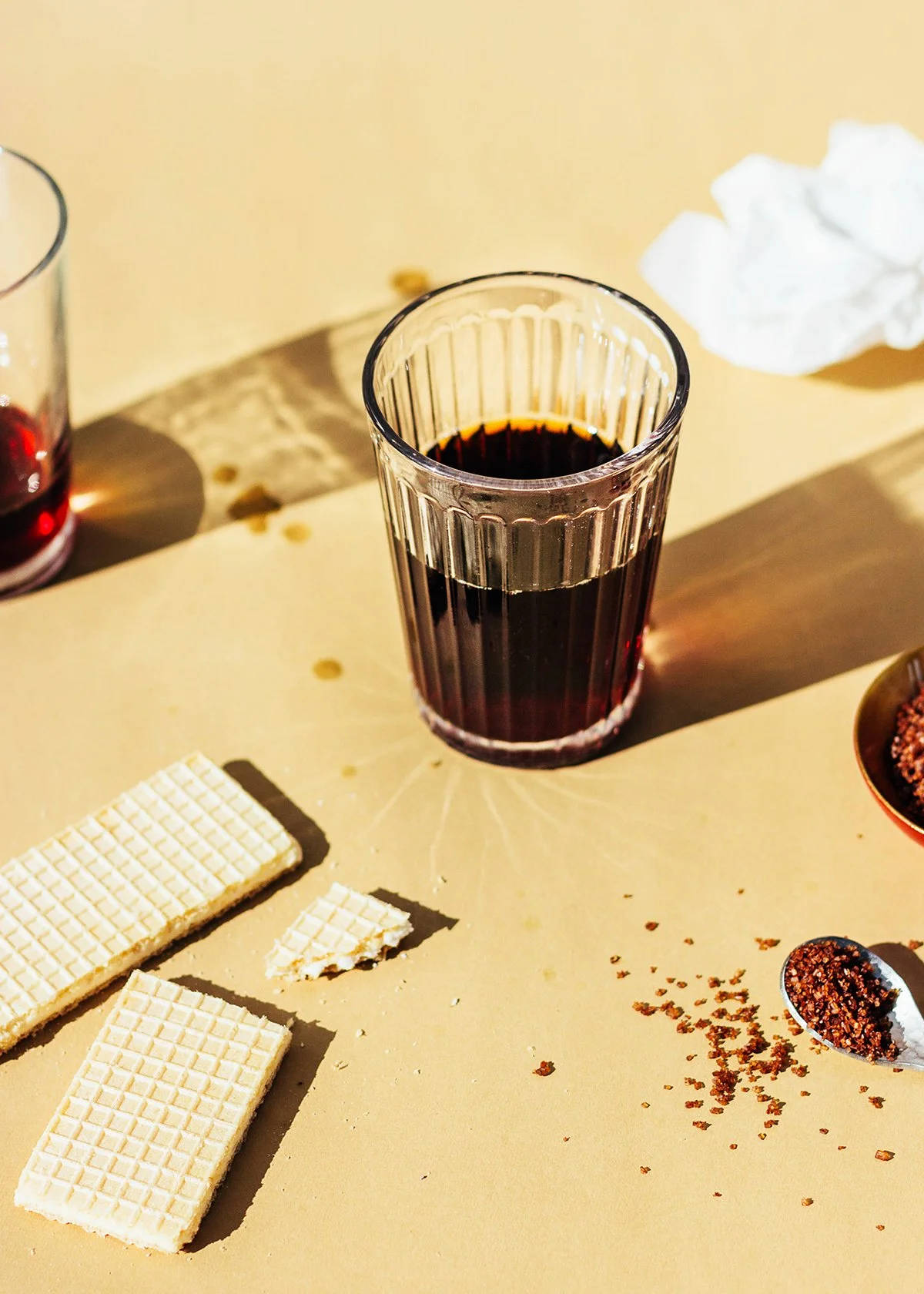 Vaso con vino tinto, tabletas de chocolate blanco, cucharas con polvo de cacao y papel arrugado sobre una mesa con manchas.
