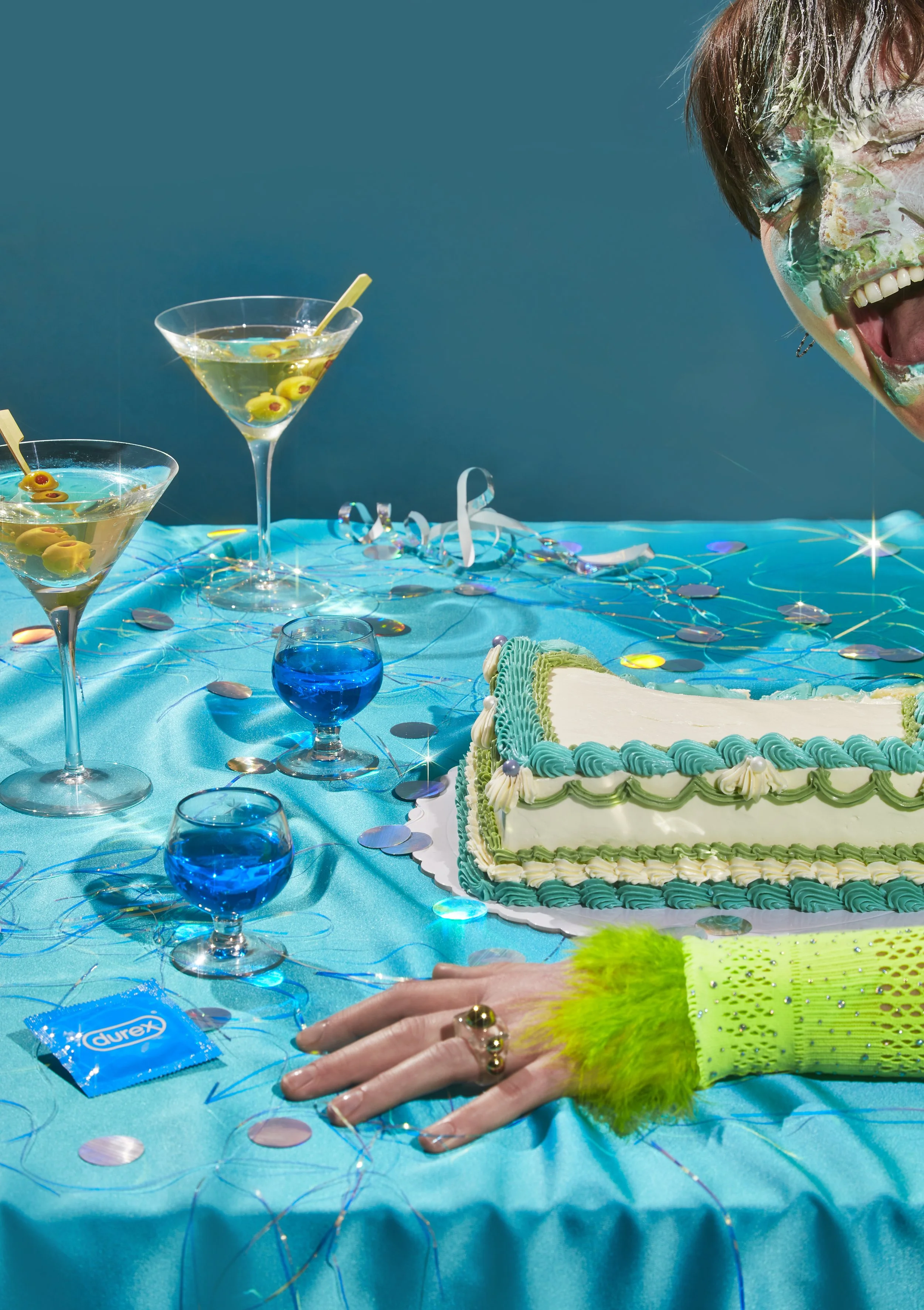 Una mesa decorada con un pastel de cumpleaños, copas con bebidas y confeti, y una persona con maquillaje colorido, vestida con tela brillante y accesorios llamativos. La celebración parece alegre y festiva.
