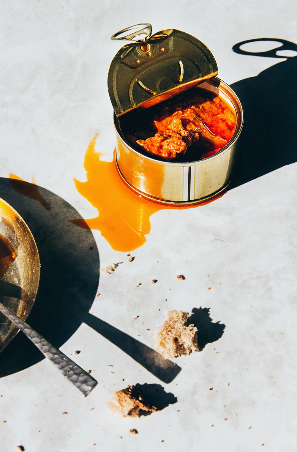Latas de comida de conservas abiertas con líquido derramado en una superficie blanca, acompañadas de una cuchara y pedazos de pan o galleta