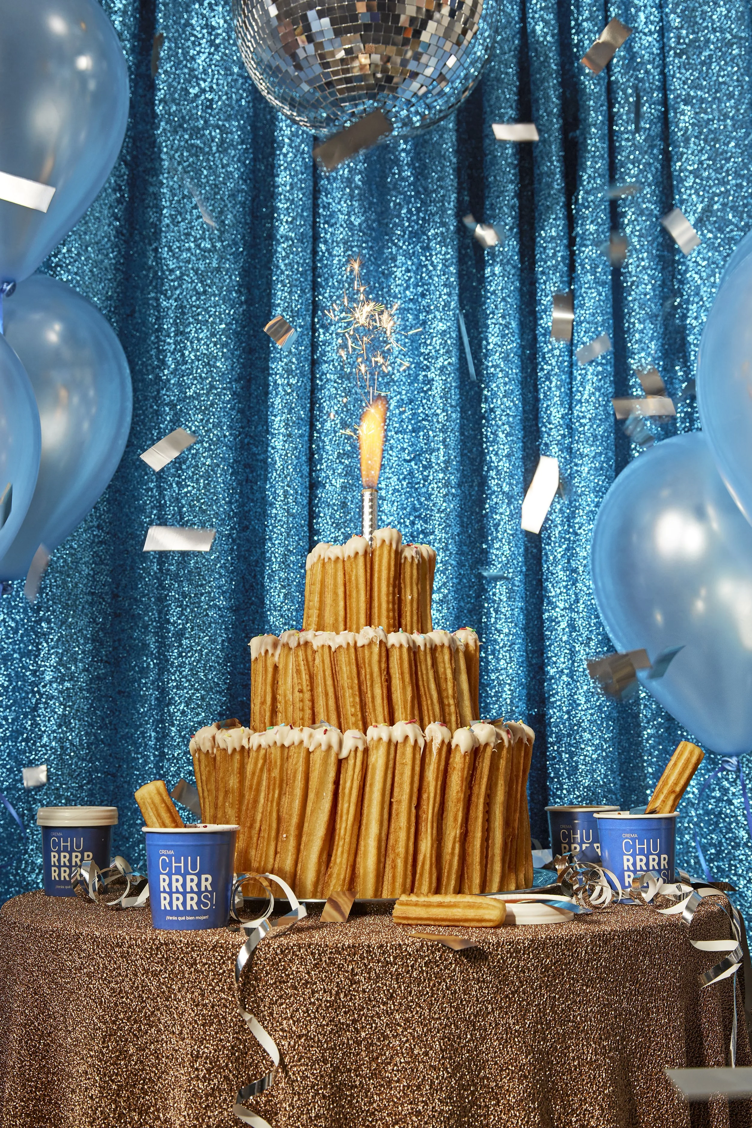 Un pastel de churros decorativos en un ambiente de fiesta con fondo de cortina azul brillante, globos azules y guirnaldas con serpentinas y un espejo de bola, con un encendedor y una chispa en la parte superior del pastel.