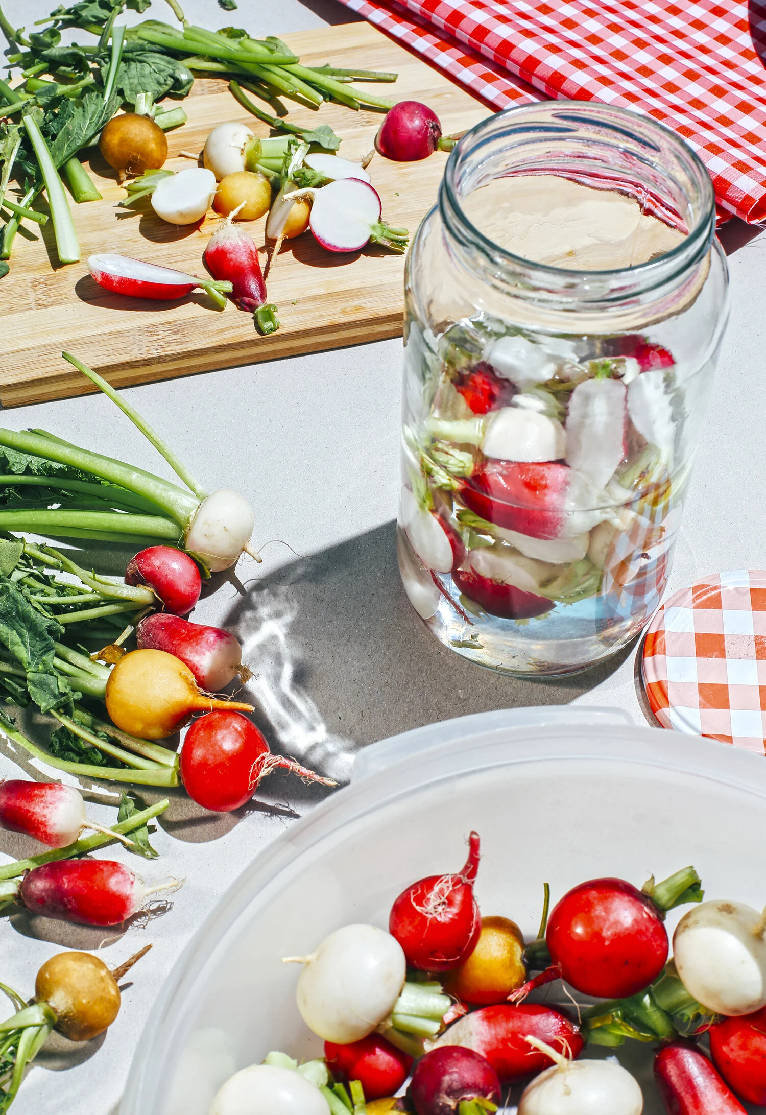 Una tabla con diferentes rábanos de varios colores, un frasco con rábanos en agua, y un recipiente con rábanos listos para preparar conservas o encurtidos.
