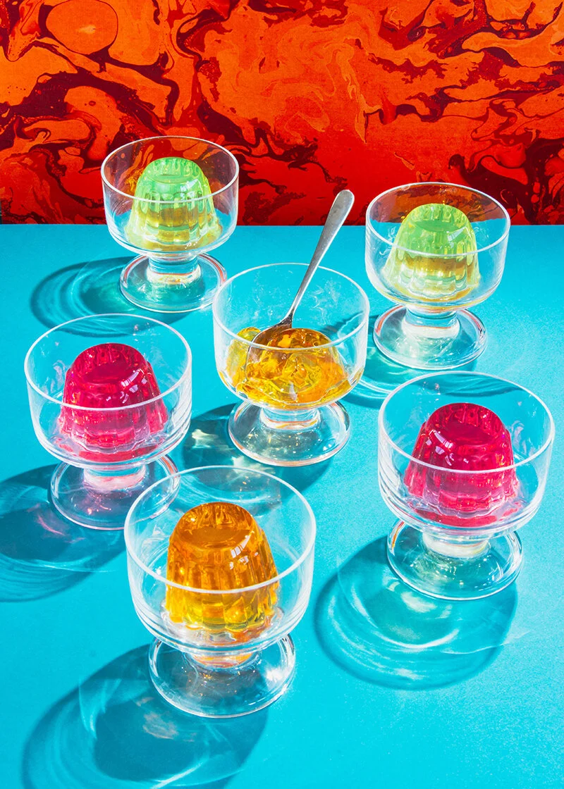 Seis copas de cristal transparente, con gelatinas de colores (roja, amarilla y verde) , en una mesa de color turquesa y fondo con patrón rojo y negro.