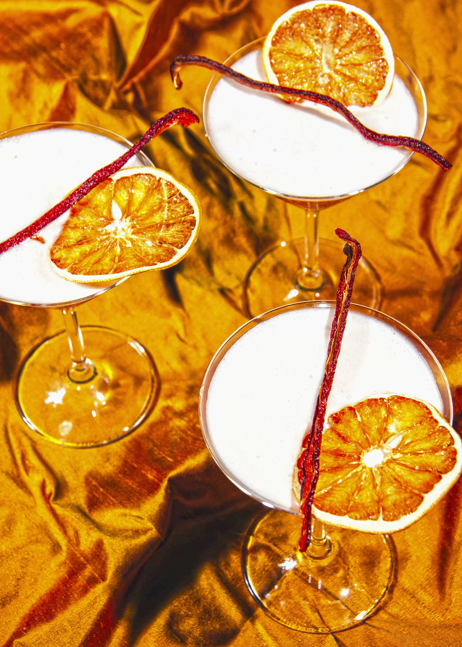 Tres copas de cóctel con decoraciones de rodajas de naranja y ramitas de canela, sobre fondo de tela dorada