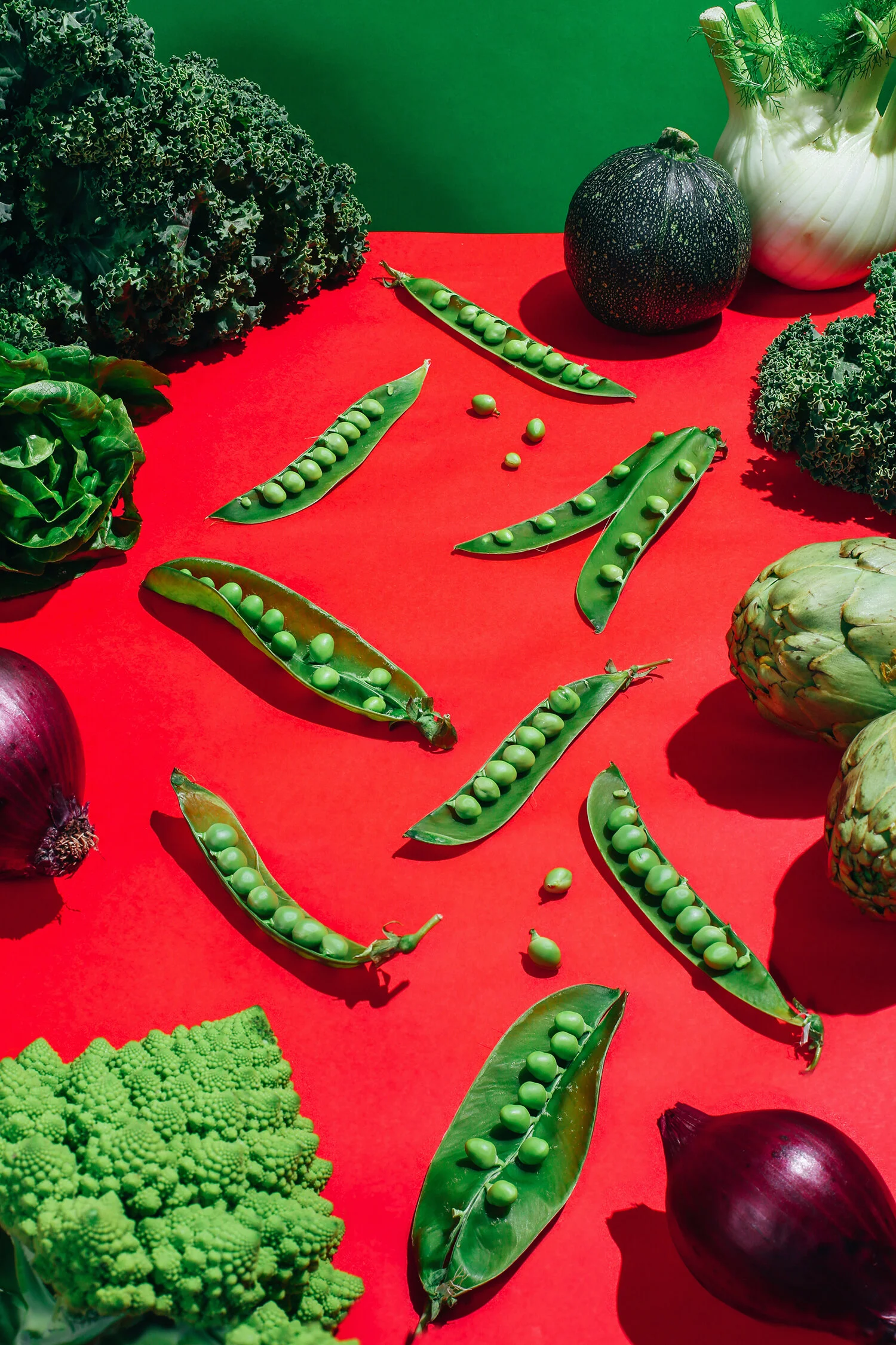 Varios tipos de verduras en una mesa roja, incluyendo guisantes en vainas abiertas, coliflor, cebolla morada, brócoli, achicoria y calabaza