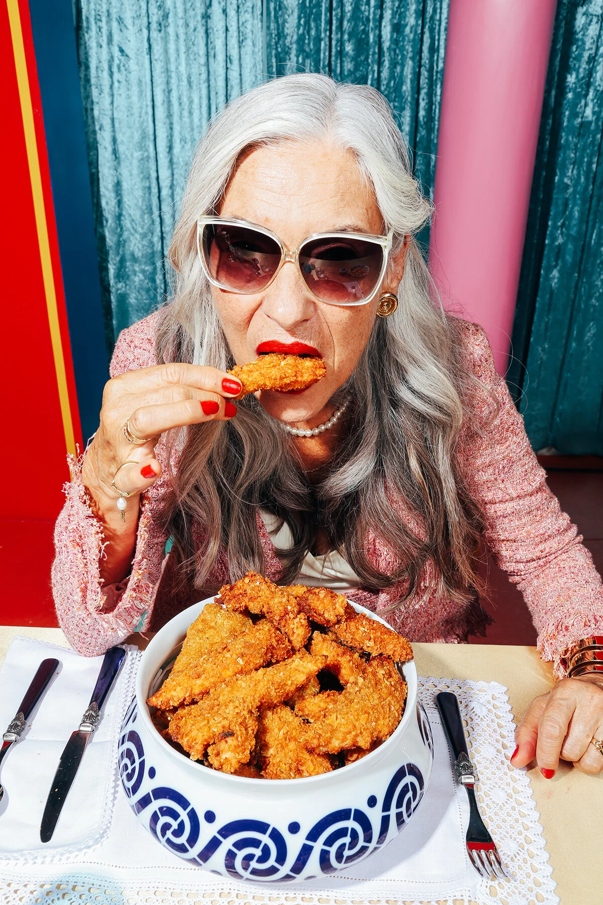 Una mujer mayor con cabello gris, gafas grandes y vestimenta elegante comiendo un trozo de pollo frito. En la mesa hay un plato grande lleno de pollo frito.