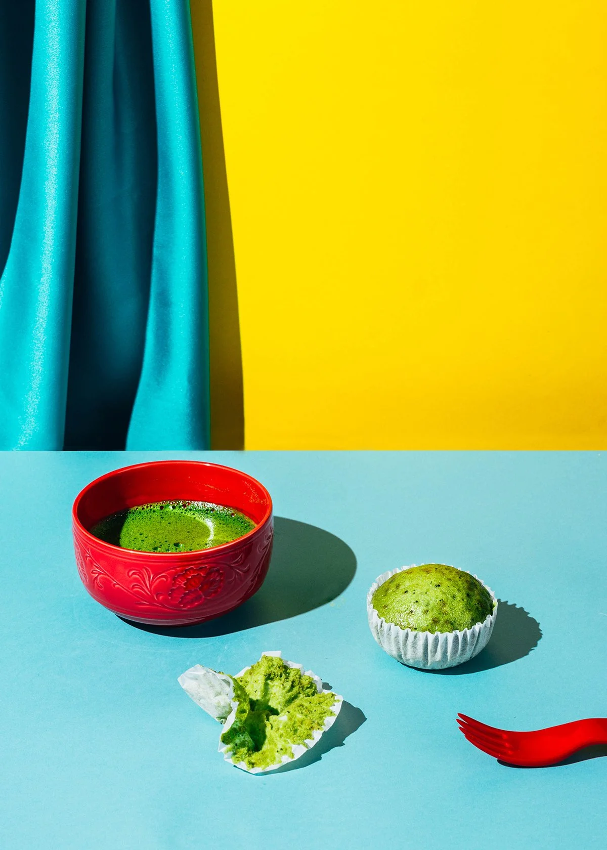 Un tazón rojo con té verde, una cupcake verde, un pedazo de cupcake y un tenedor rojo sobre un fondo de mesa azul, con cortinas azules y pared amarilla en el fondo.