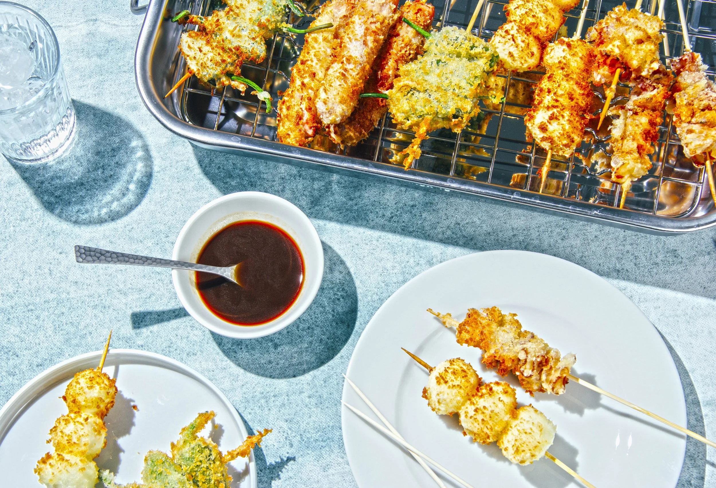 Brochetas de alimentos empanizados y fritos, servidas en plato y en bandeja, acompañadas de salsa oscura, en una mesa con vaso de agua.