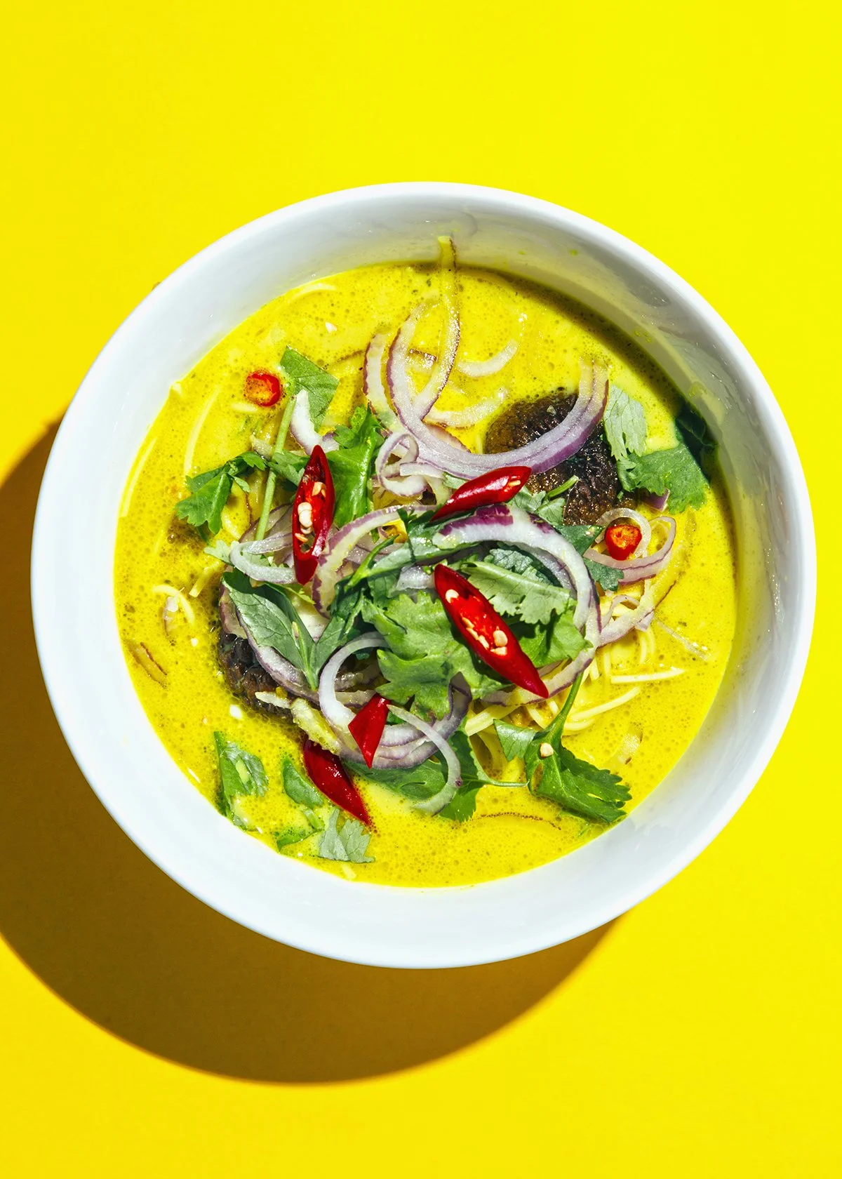 Curry tailandés con cebolla roja, cilantro y chiles rojos en un bol blanco sobre fondo amarillo.