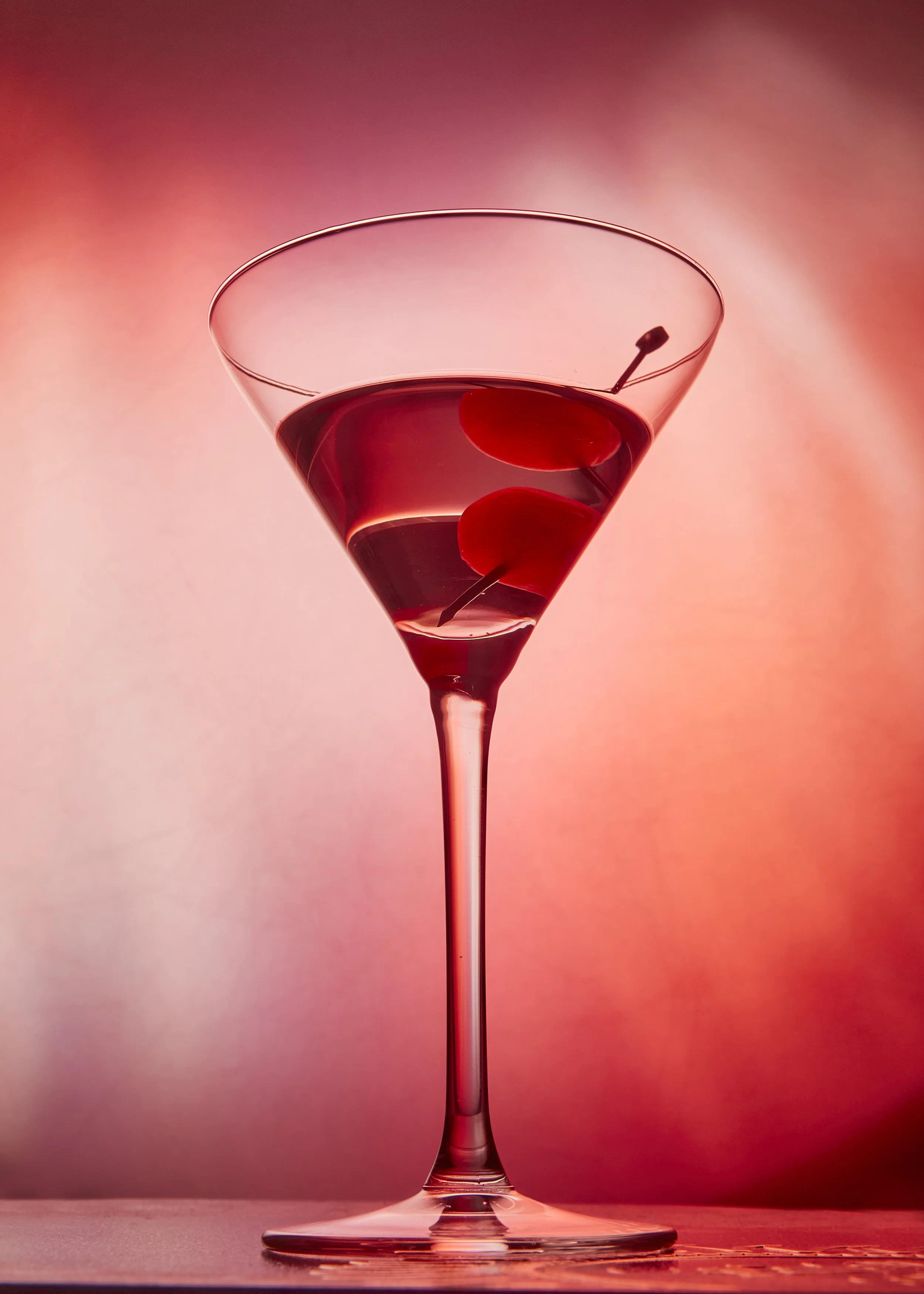 Vaso de cóctel con aceitunas en un fondo difuso de tonos rosados y rojos.