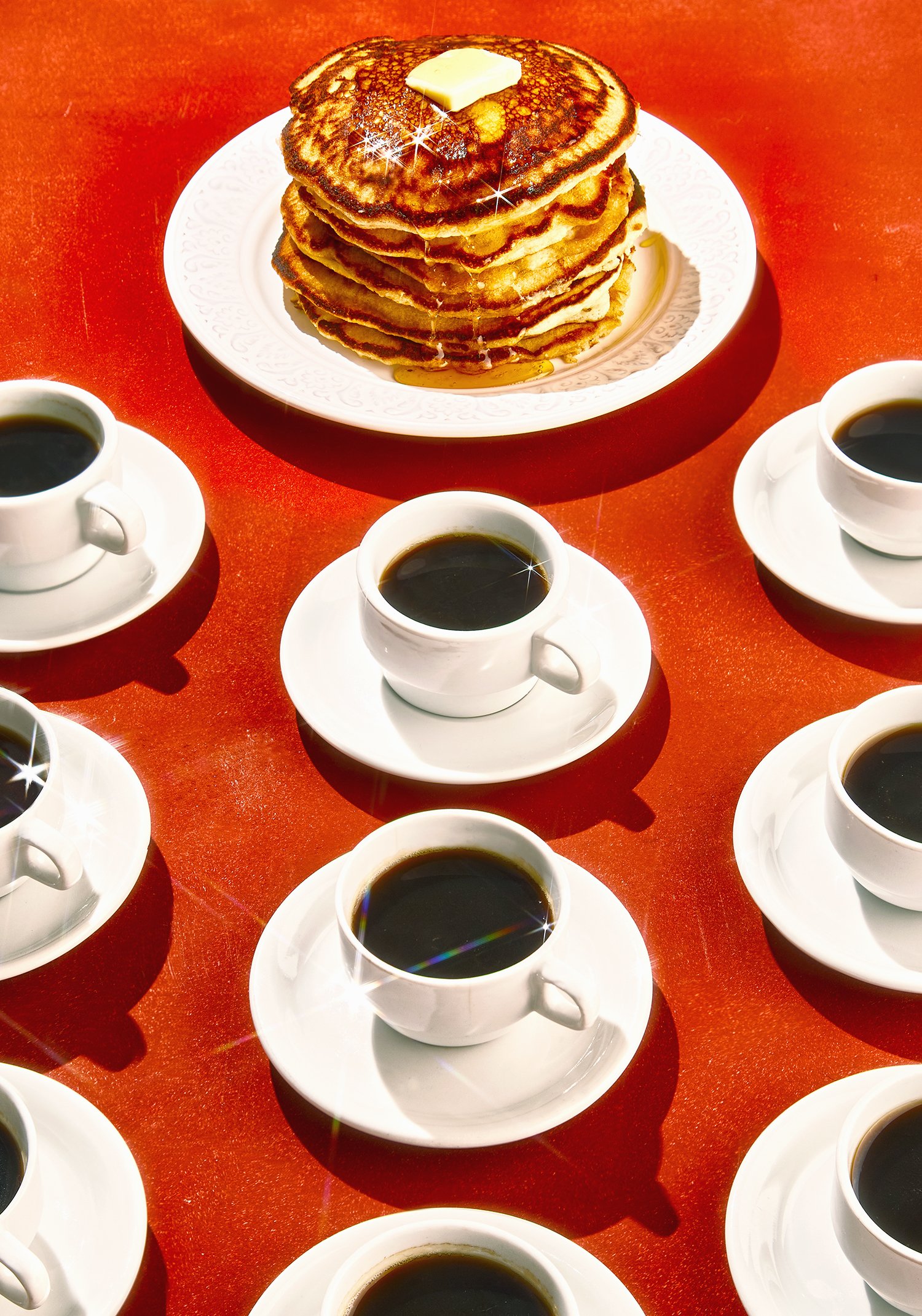 Un plato con varias tortitas apiladas, con mantequilla y jarabe en una mesa roja, rodeado de tazas de café.