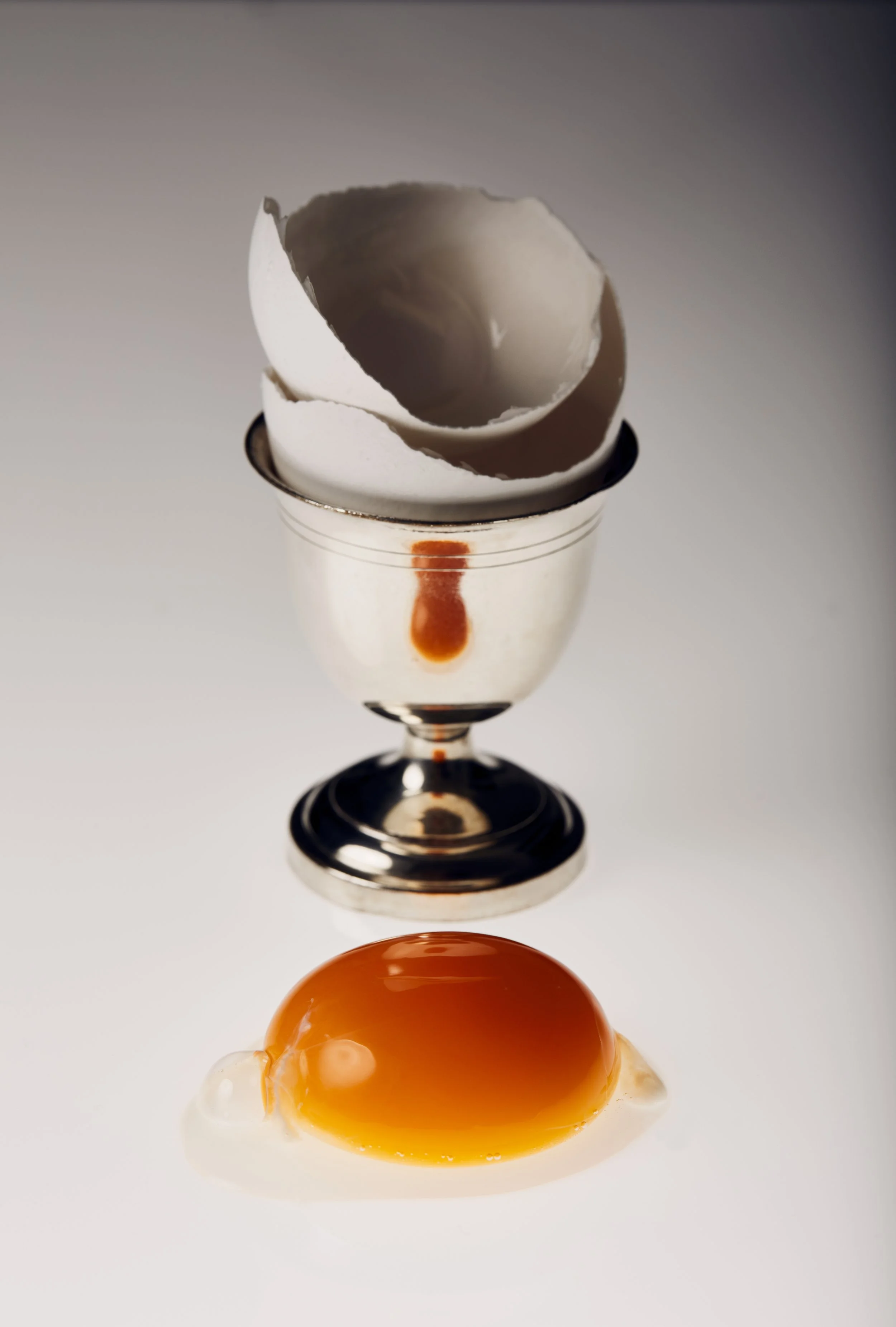 Huevo crudo con yema líquida en primer plano, y cascarón de huevo roto sobre una copa de metal en el fondo.