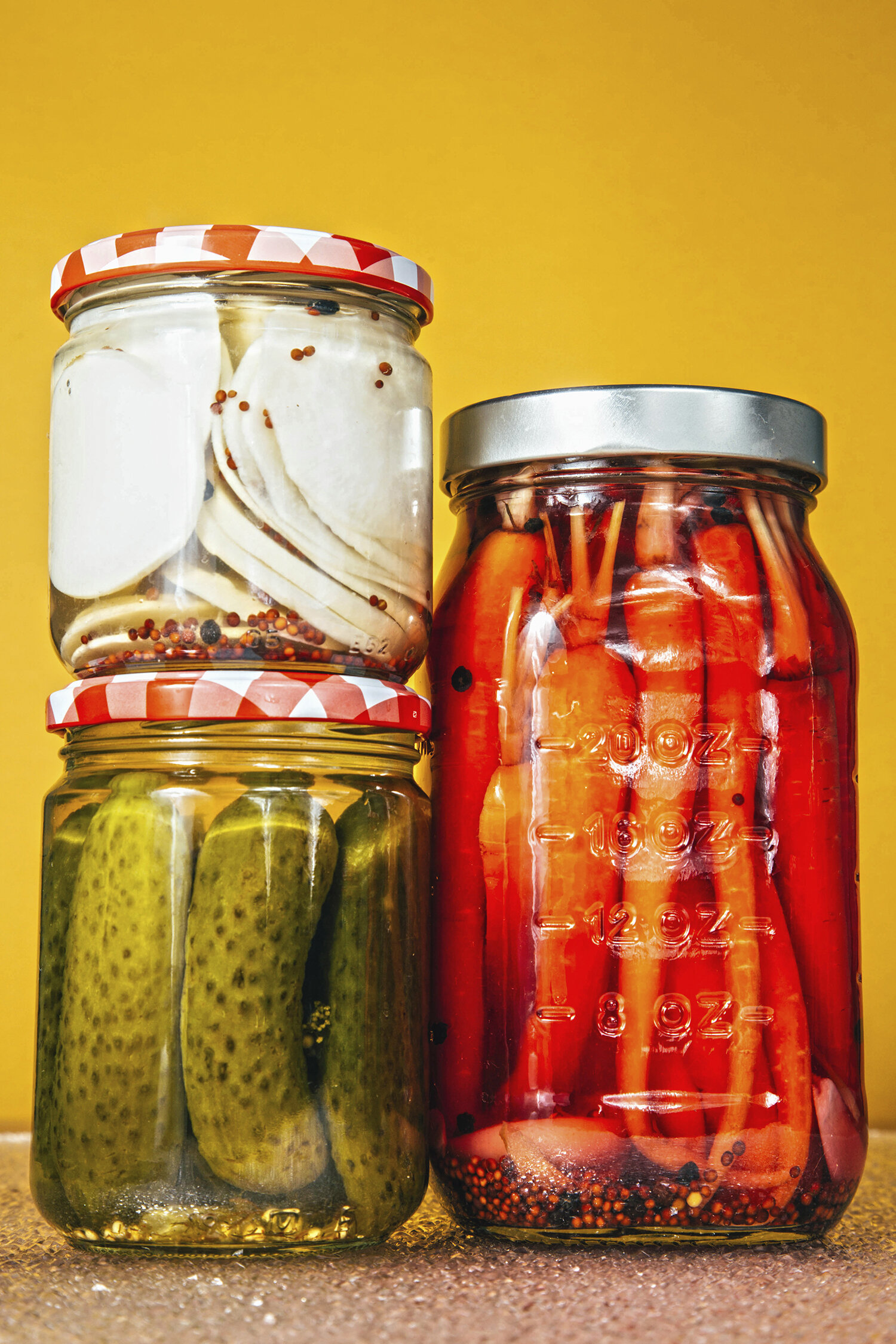 Tres frascos de conservas con pepinos, pimientos y otros vegetales en vinagre y especias, sobre fondo amarillo.
