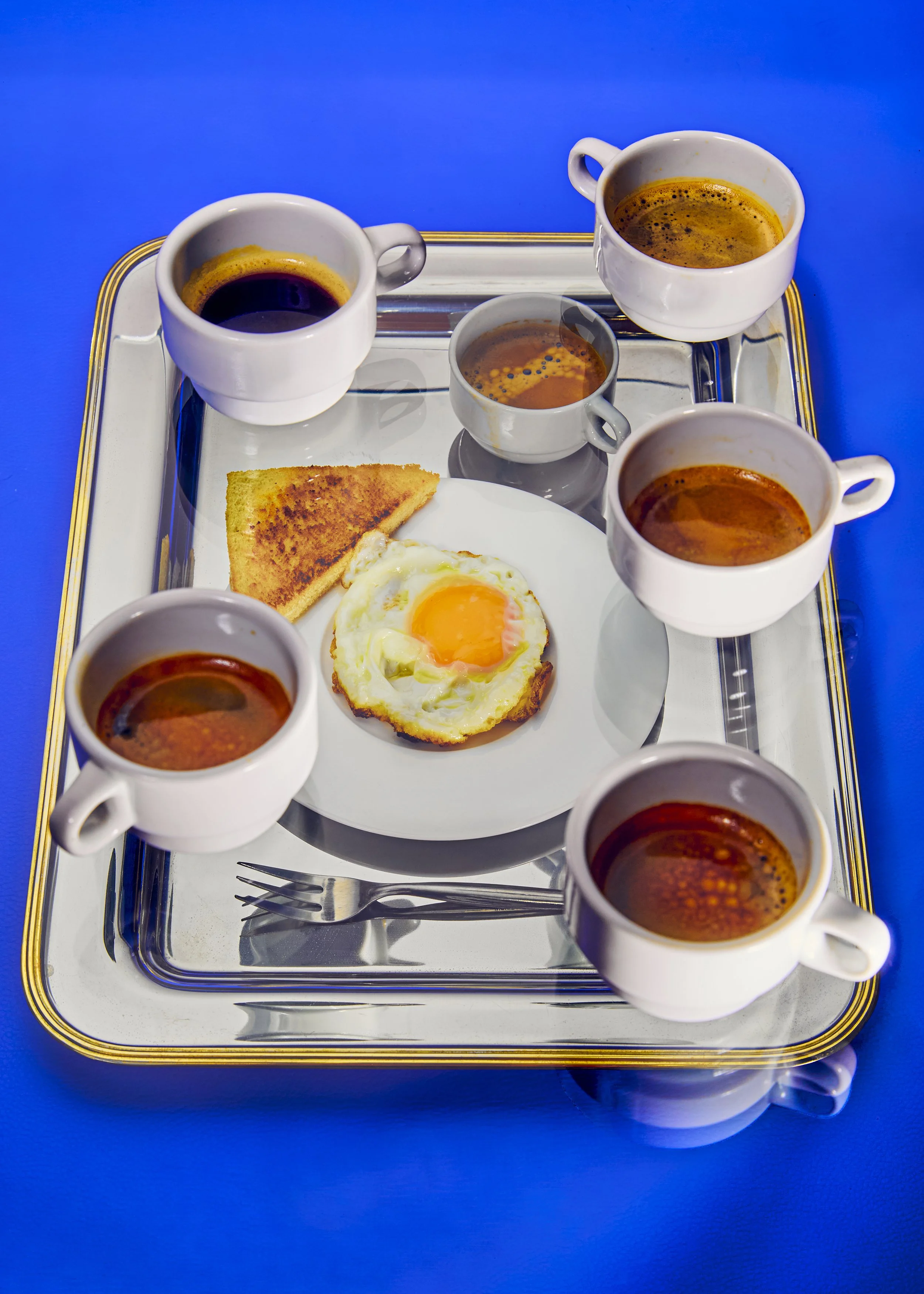 Desayuno con huevos fritos, rebanada de pan tostado, seis tazas de café en platos blancos sobre una bandeja plateada.