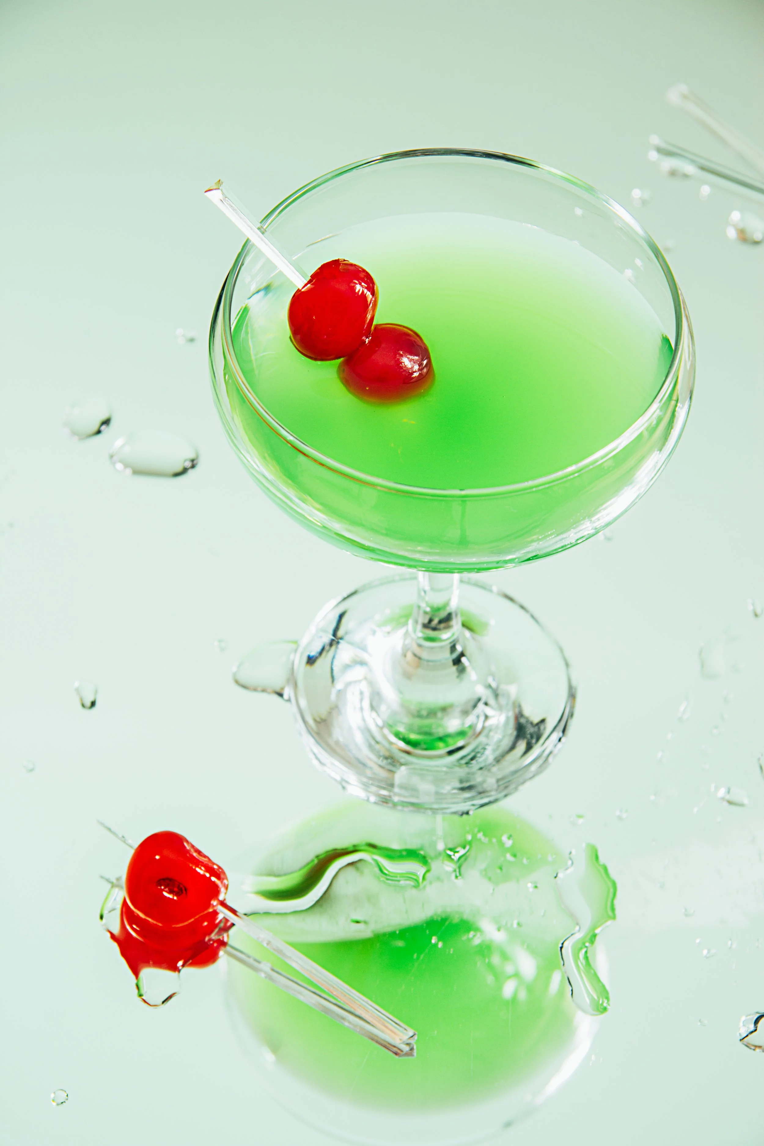 Copa de cóctel verde con cerezas y una cereza en una brocheta, con gotas de agua alrededor.